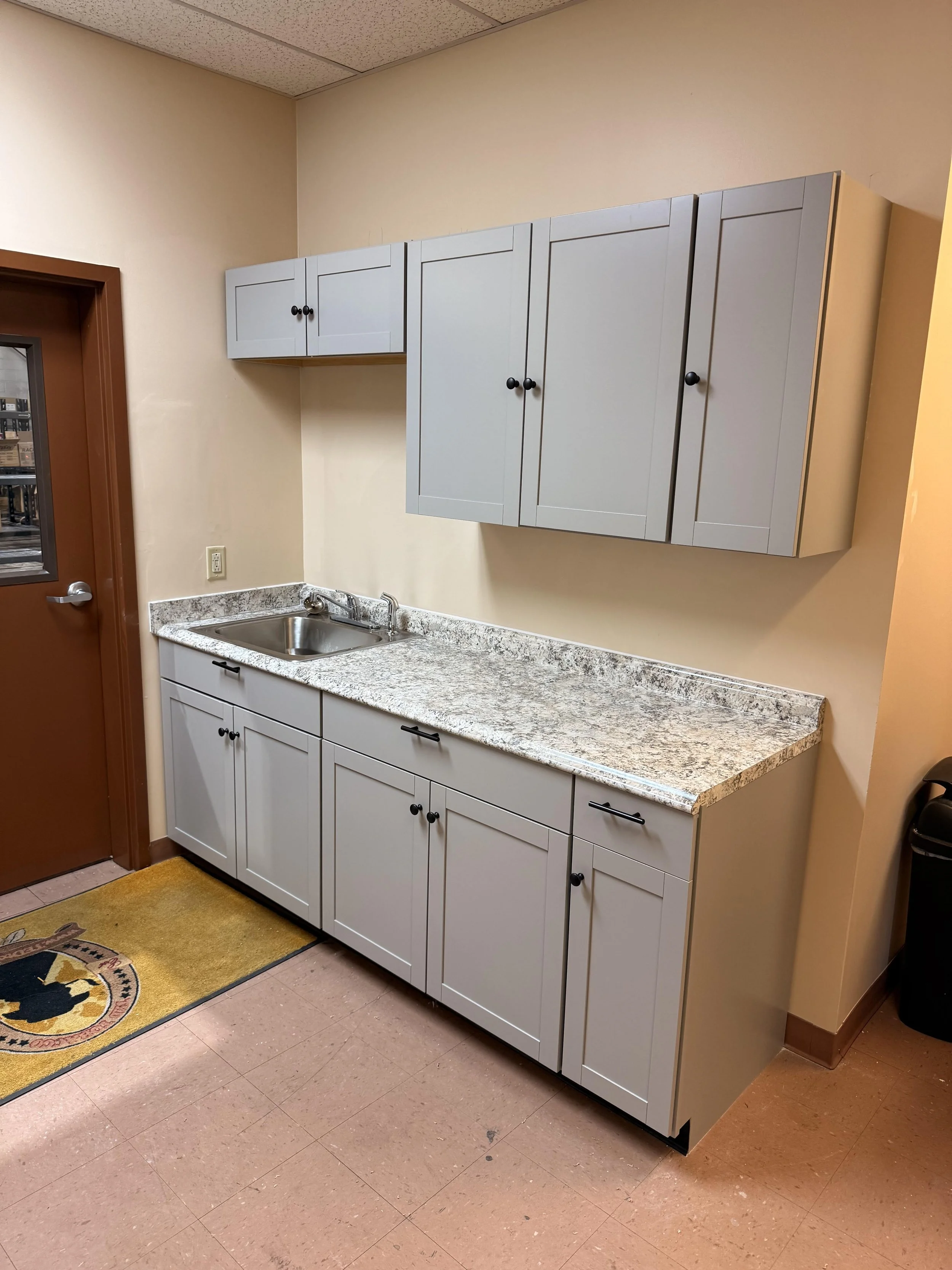 A kitchen or utility area with white cabinets mounted on a beige wall, a granite countertop, and a stainless steel sink. There is a door on the left side with a window and a decorative mat on the floor.