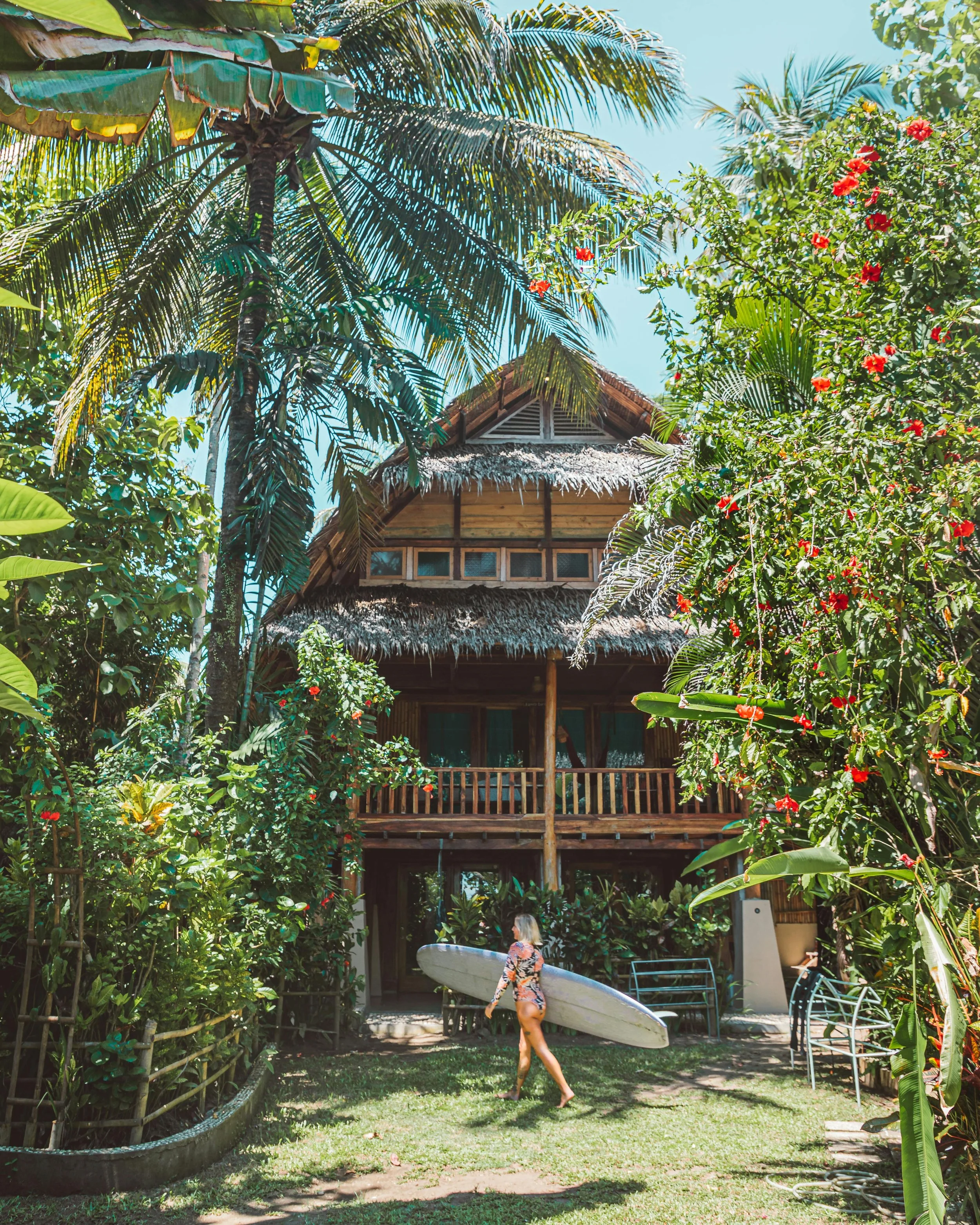 A woman in a floral swimsuit holding a surfboard walks on a grassy area near a wooden tropical-style house surrounded by lush green plants and tall palm trees.