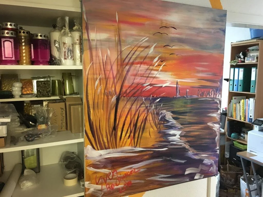 Ein Raum mit Regalen, die Gläser und Kerzenhalter enthalten. Daneben ein großes, buntes Gemälde, das einen Sonnenuntergang über einem Gewässer mit Vögeln am Himmel darstellt. Im Hintergrund sind weitere Regale mit Ordnern und Büchern zu sehen.