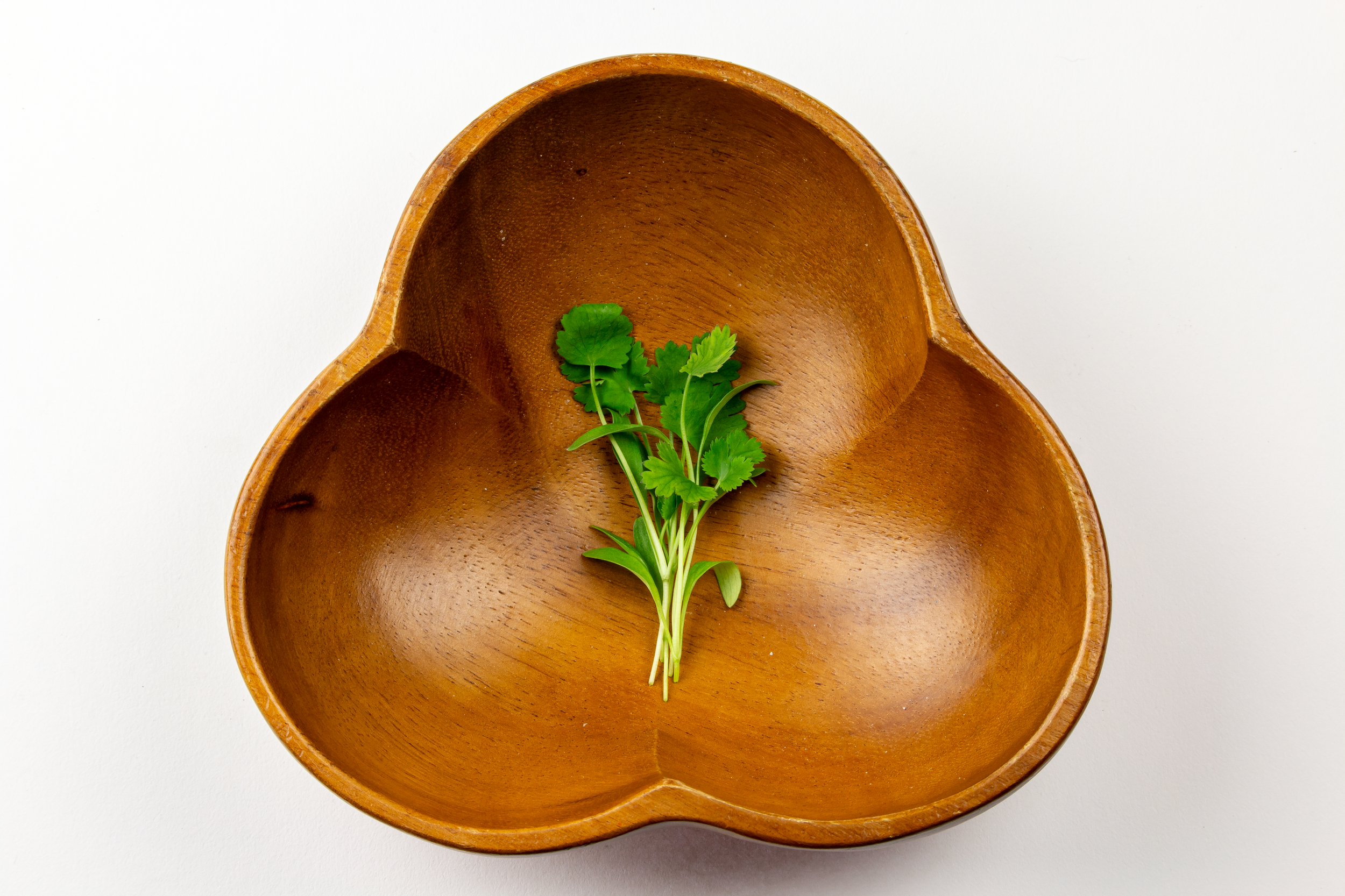A wooden heart-shaped bowl with a small bunch of fresh green herbs inside on a white surface.
