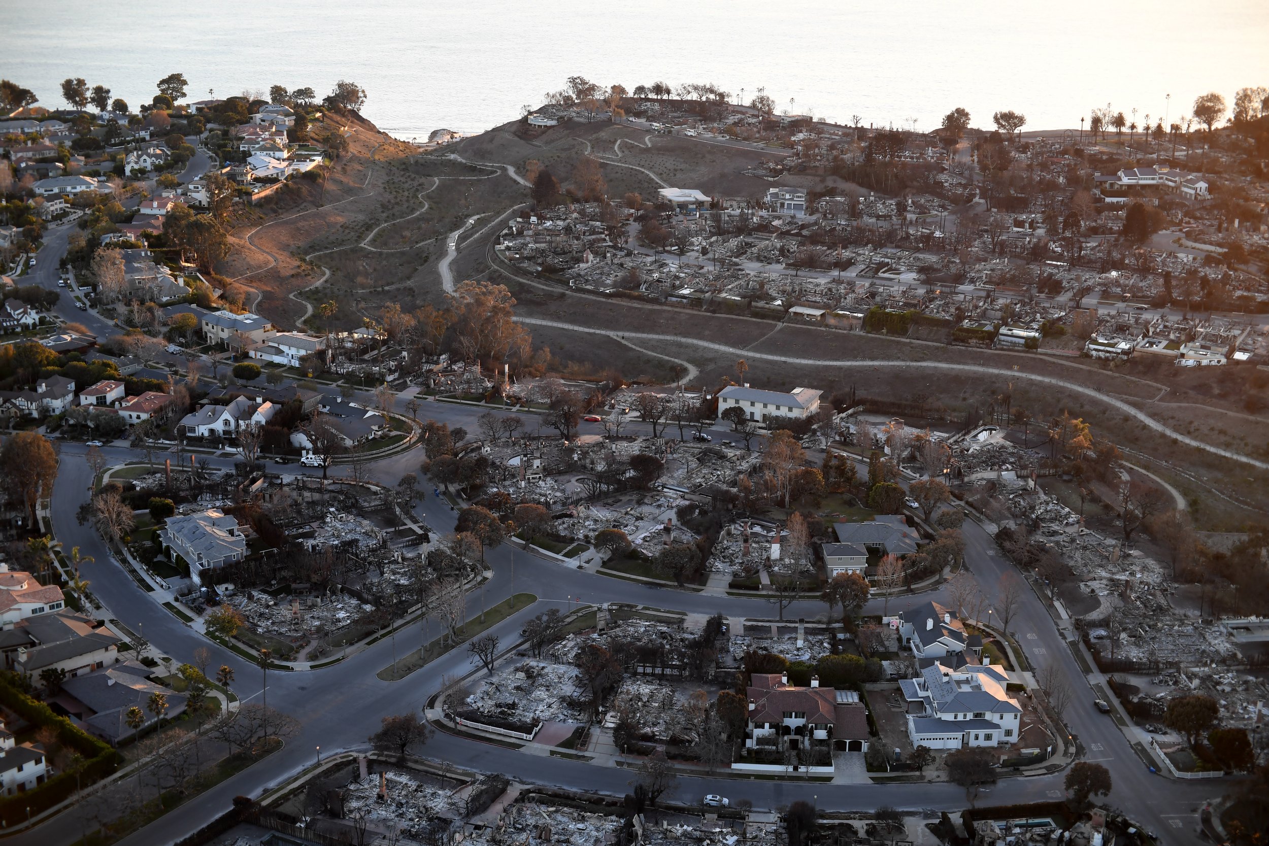 Aerial view of a neighborhood partially destroyed by a wildfire, with some houses intact and others reduced to rubble and ash.