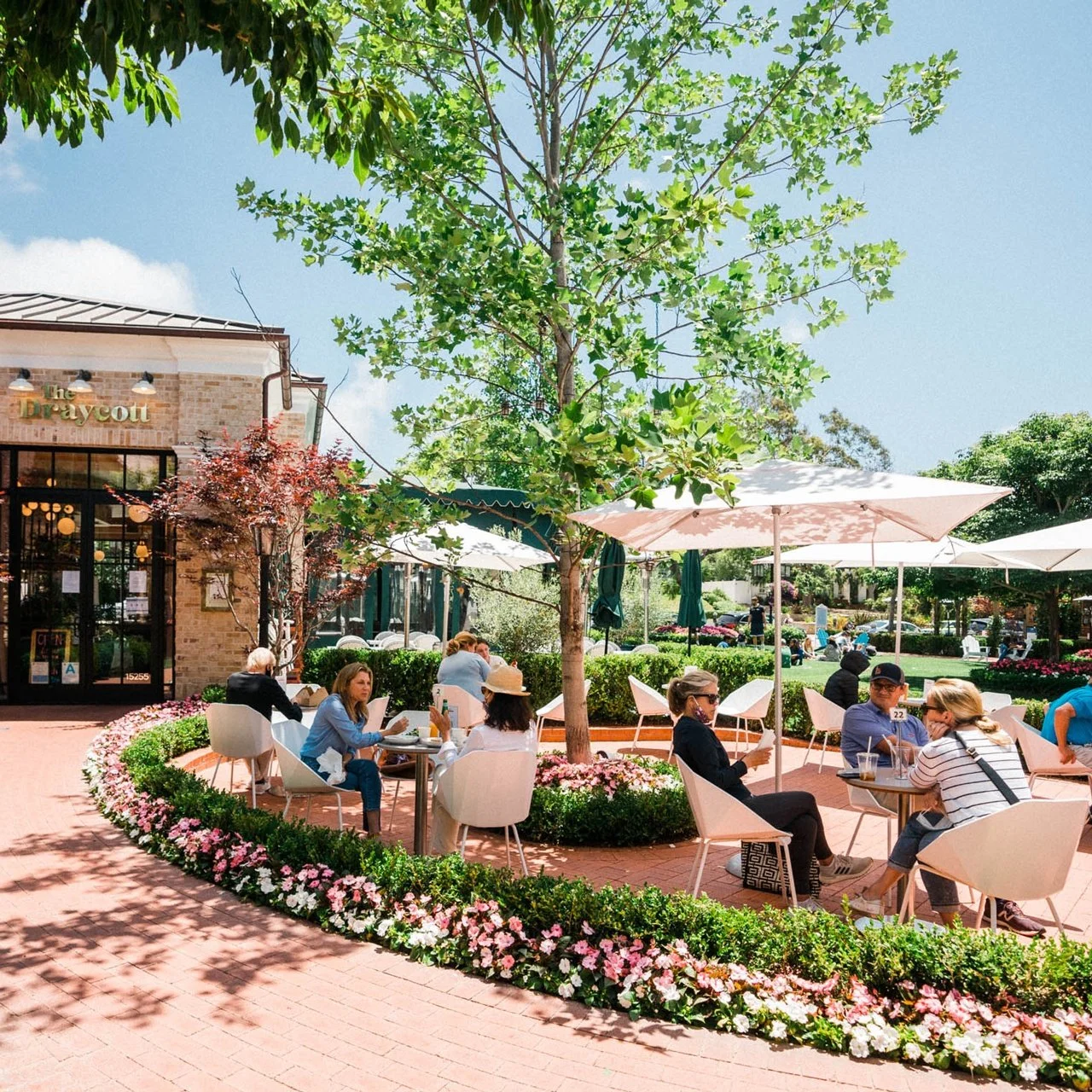 Outdoor cafe with people dining under umbrellas, surrounded by greenery and flowers, features a brick pathway and building entrance.