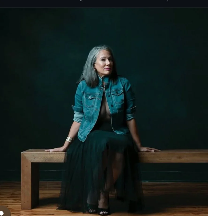 Woman in denim jacket and black skirt sitting on wooden bench against dark background.
