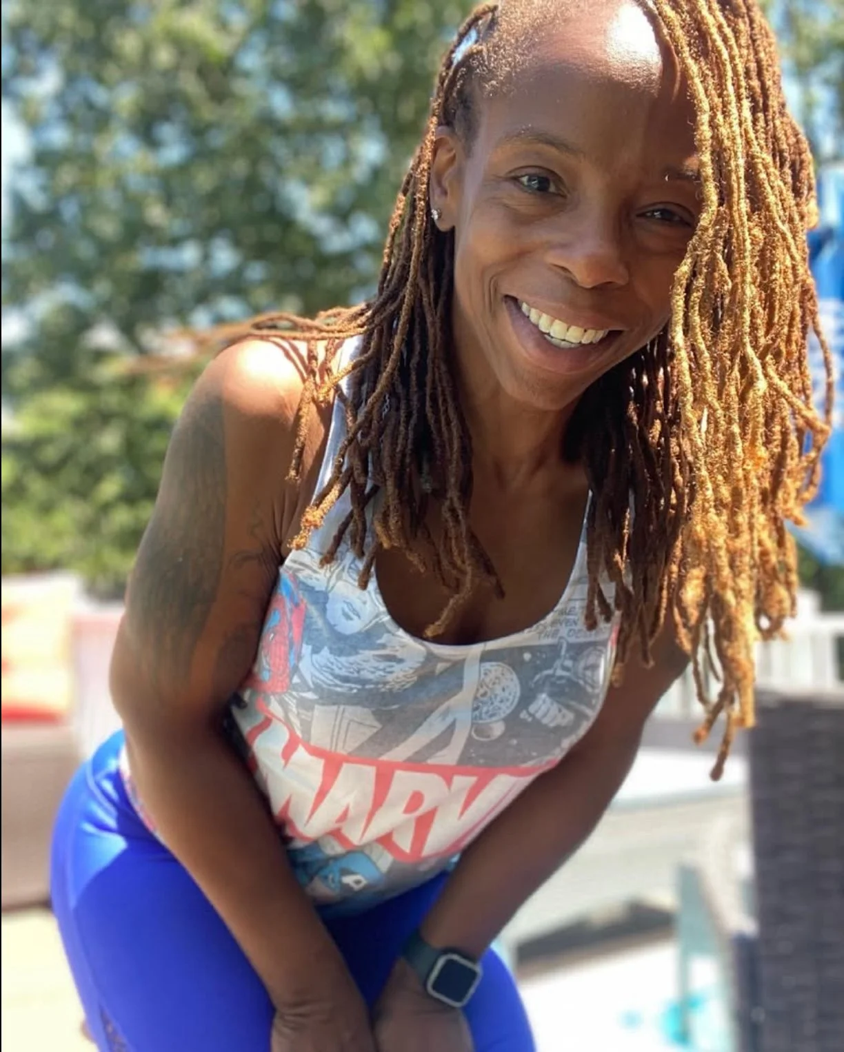Smiling person with long dreadlocks wearing a Marvel tank top and blue pants outdoors.