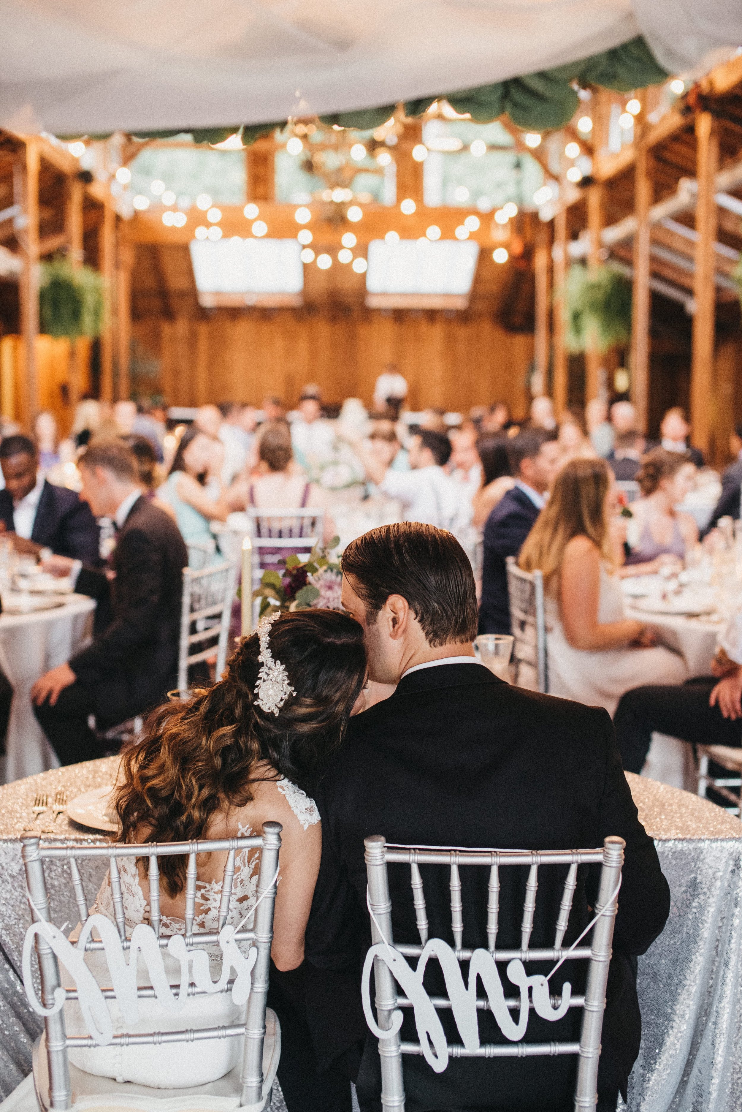 A bride and groom sitting close together, with the bride resting her head on the groom's shoulder, at their wedding reception in a rustic wooden venue with string lights overhead.