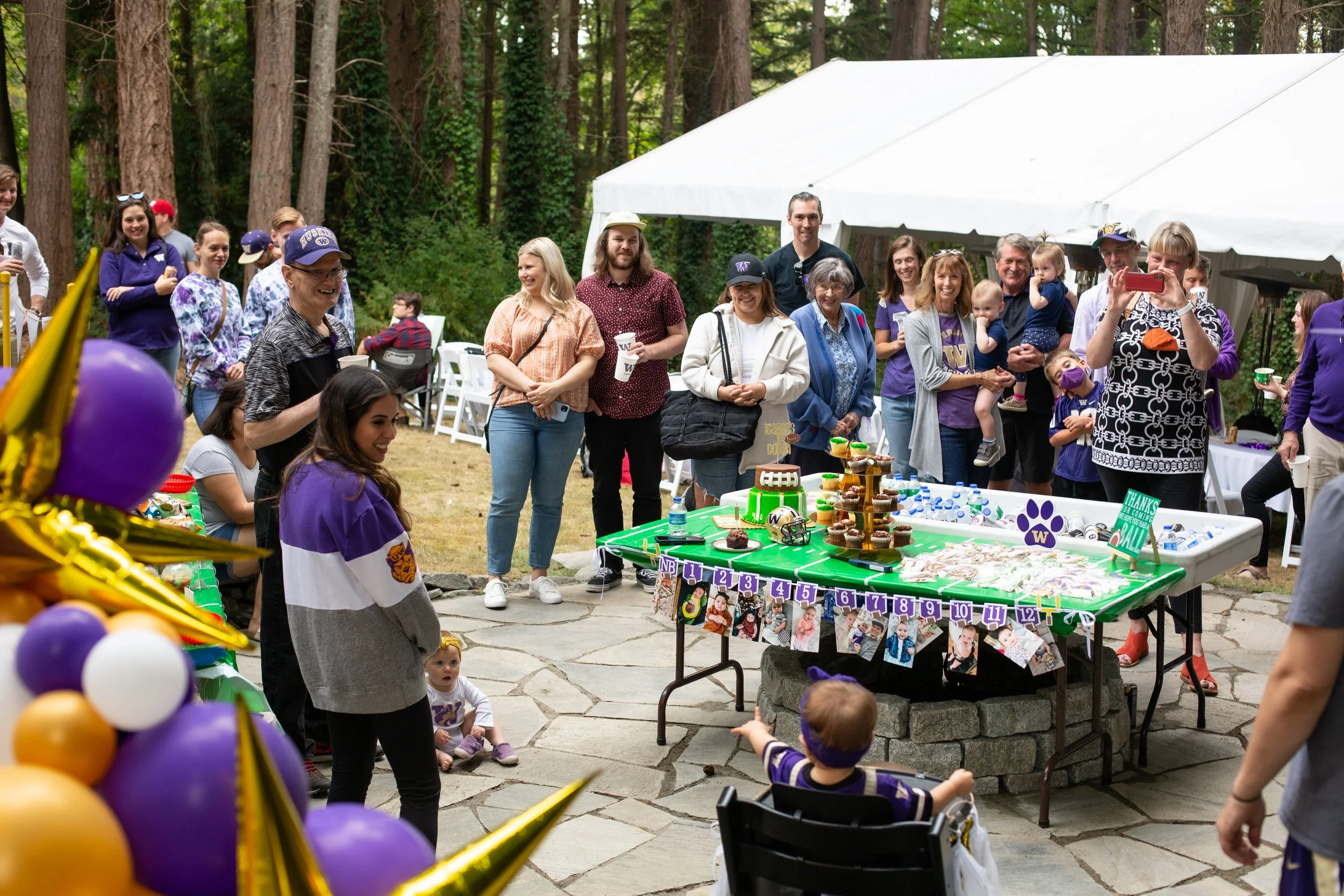 A group of people at a birthday party outdoors, with children and adults gathered around a table with cupcakes and decorations, under a white canopy in a wooded area.