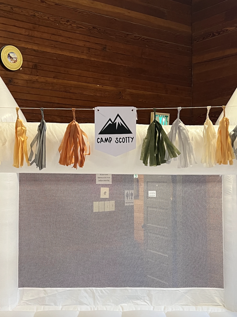 Tent decorated with multi-colored tassels and a Camp Scotty banner, inside a wooden-paneled cabin.