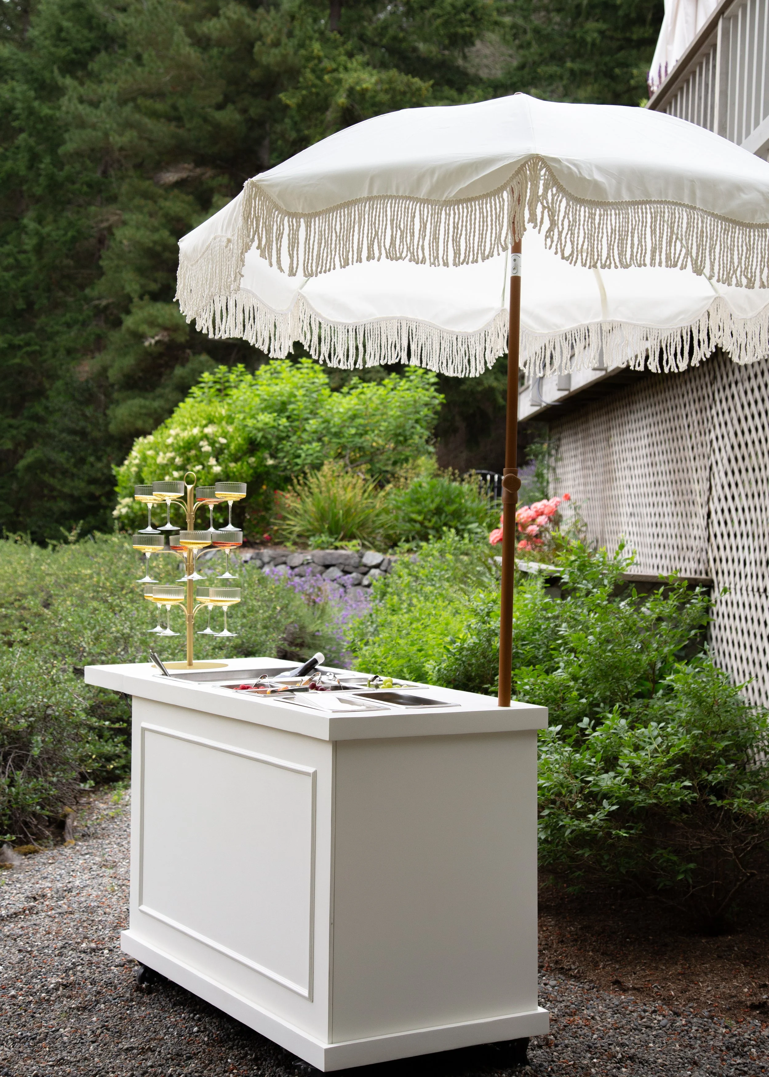 An outdoor ice cream cart with a white body and black wheels, situated on a gravel path. It includes a small section with toppings and a tiered display of glasses, shaded by a large, white, fringed umbrella. The background features lush green bushes and trees.