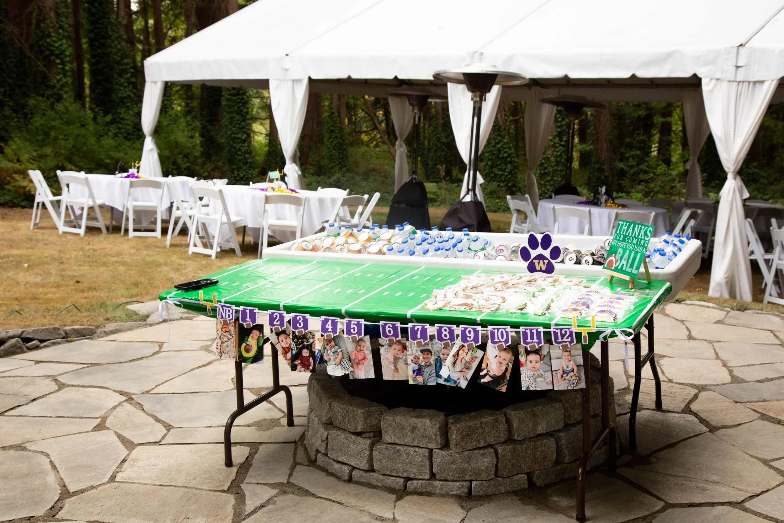 Outdoor birthday party setup with a table decorated as a football field, photos of a child hanging underneath, and a sign that says 'Thanks for coming, we hope you had a ball!'. A white canopy with tables and chairs behind, and a wooded background.