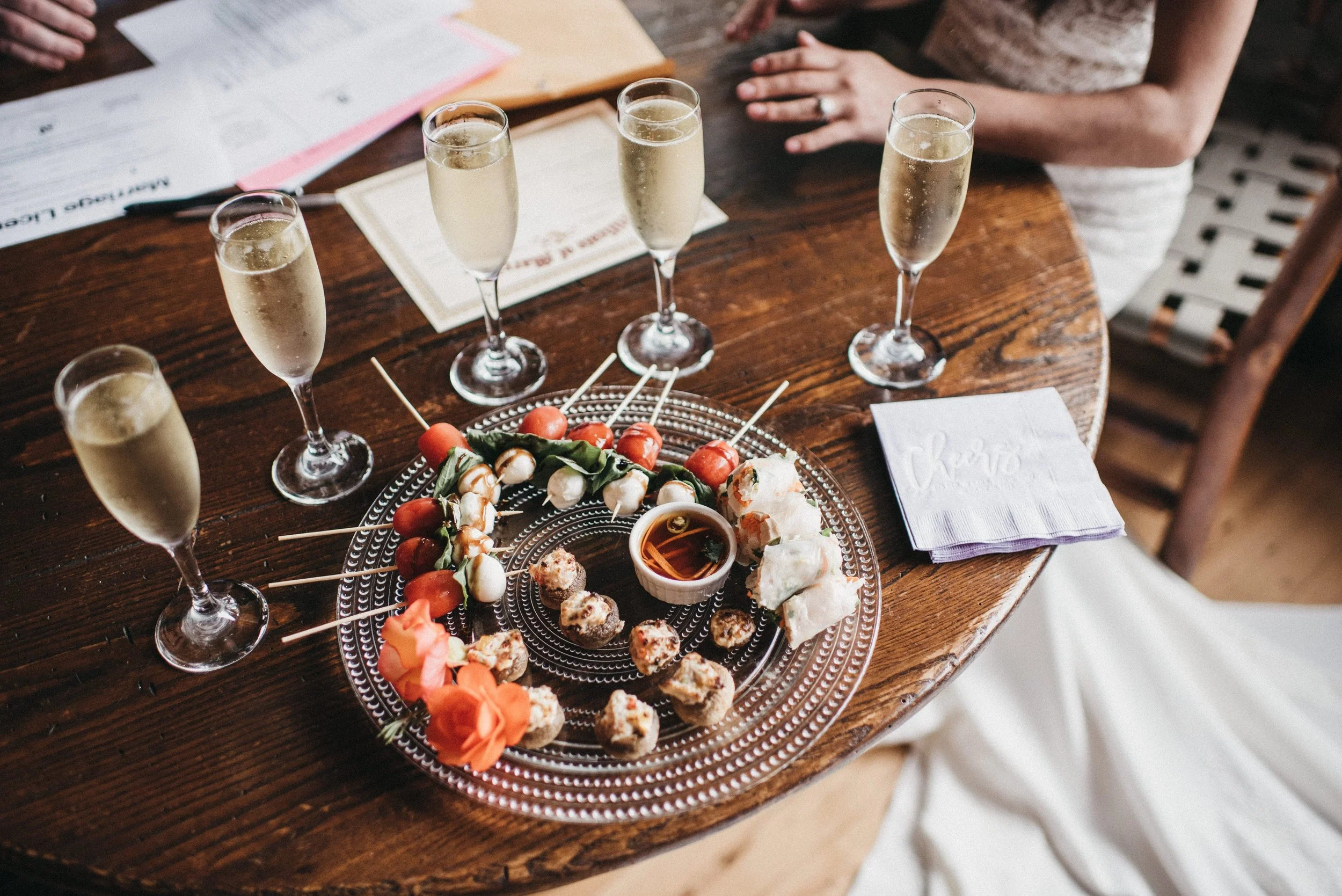 A round wooden table with six glasses of champagne, a plate of skewered appetizers, and a napkin.