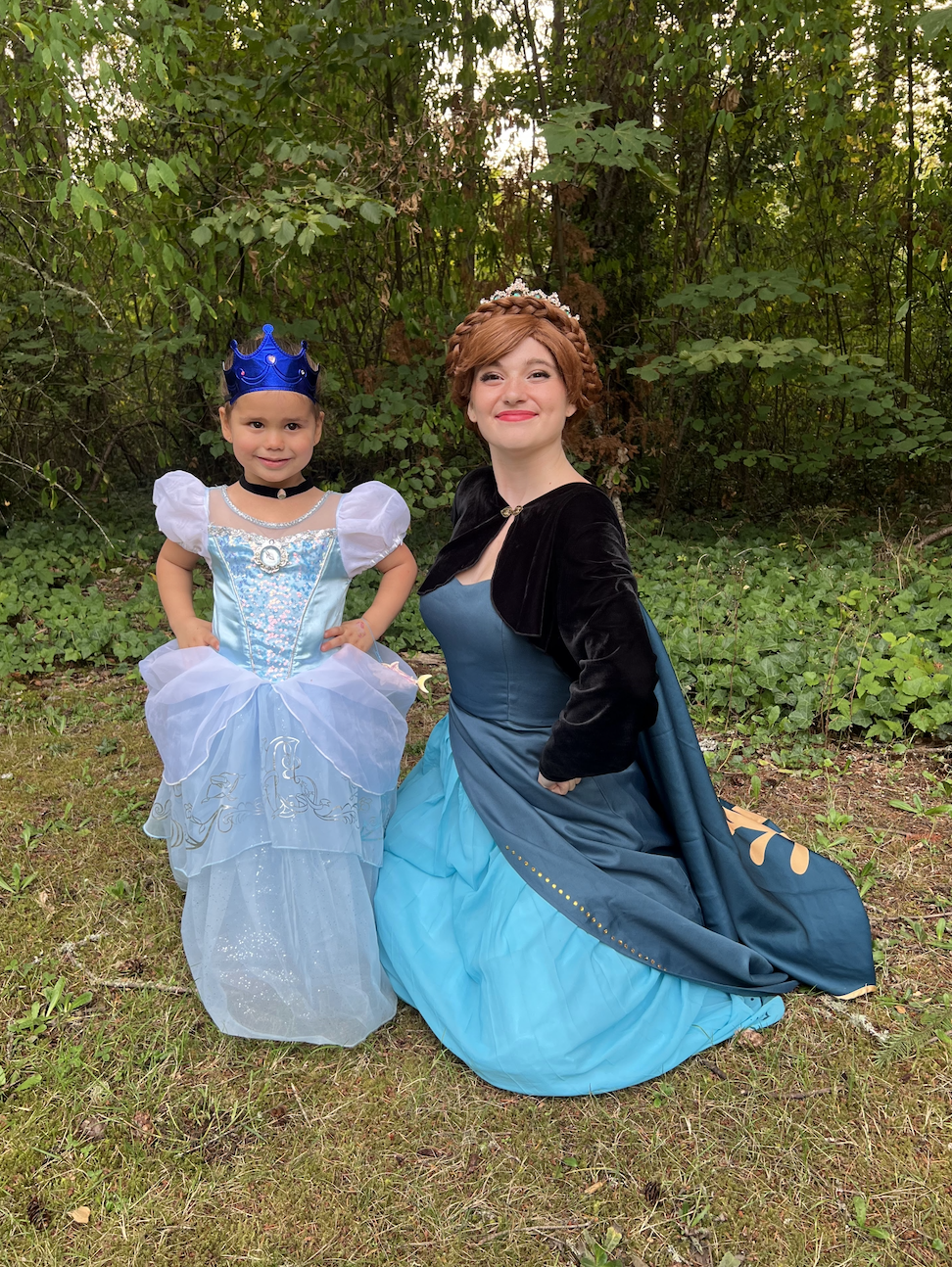 A young girl and a woman dressed as princesses at a wooded outdoor location. The girl wears a blue princess dress with white puffy sleeves and a blue crown, while the woman wears a blue gown and a black cloak, with a crown on her head. They are smili