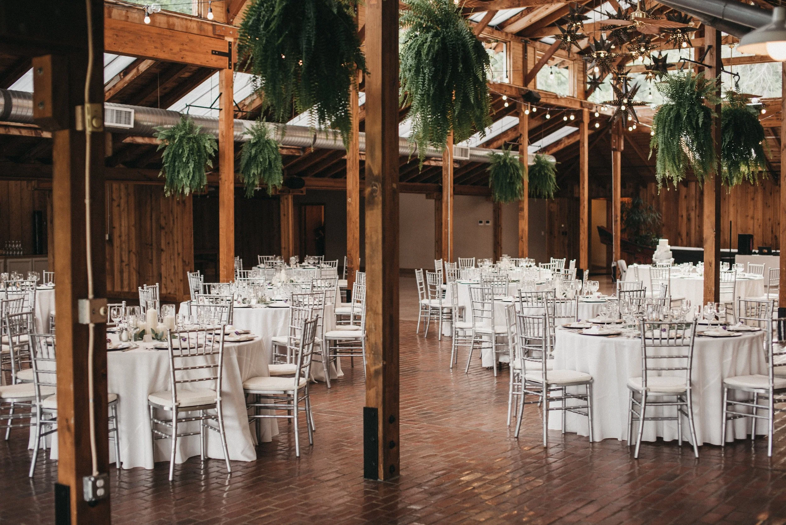 Wedding reception setup with round tables draped in white tablecloths and silver chairs, decorated with candles, in a wooden barn with green hanging ferns and star-shaped light fixtures overhead.