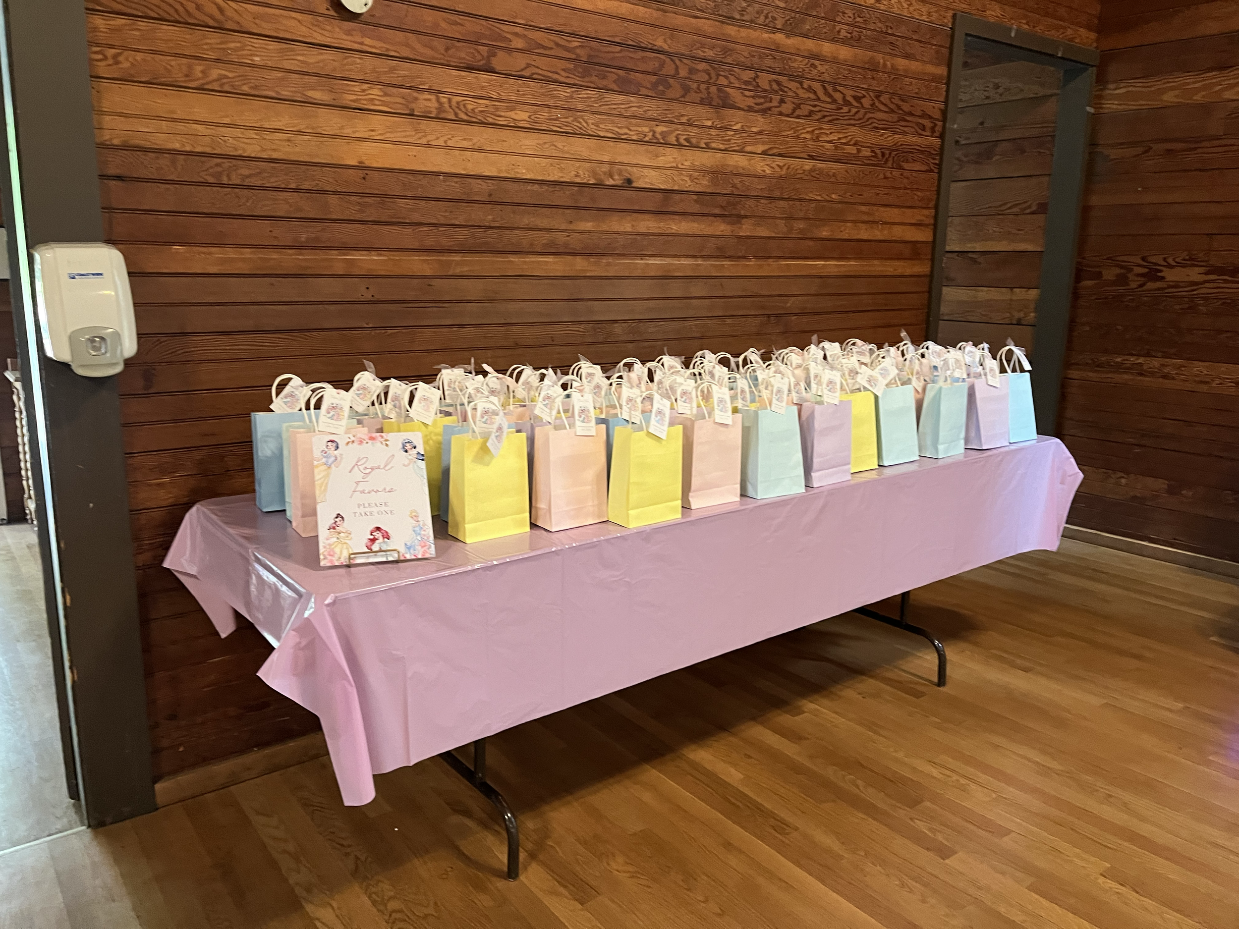 Table with pastel-colored gift bags on a pink tablecloth, set against a wooden wall at a party or event.
