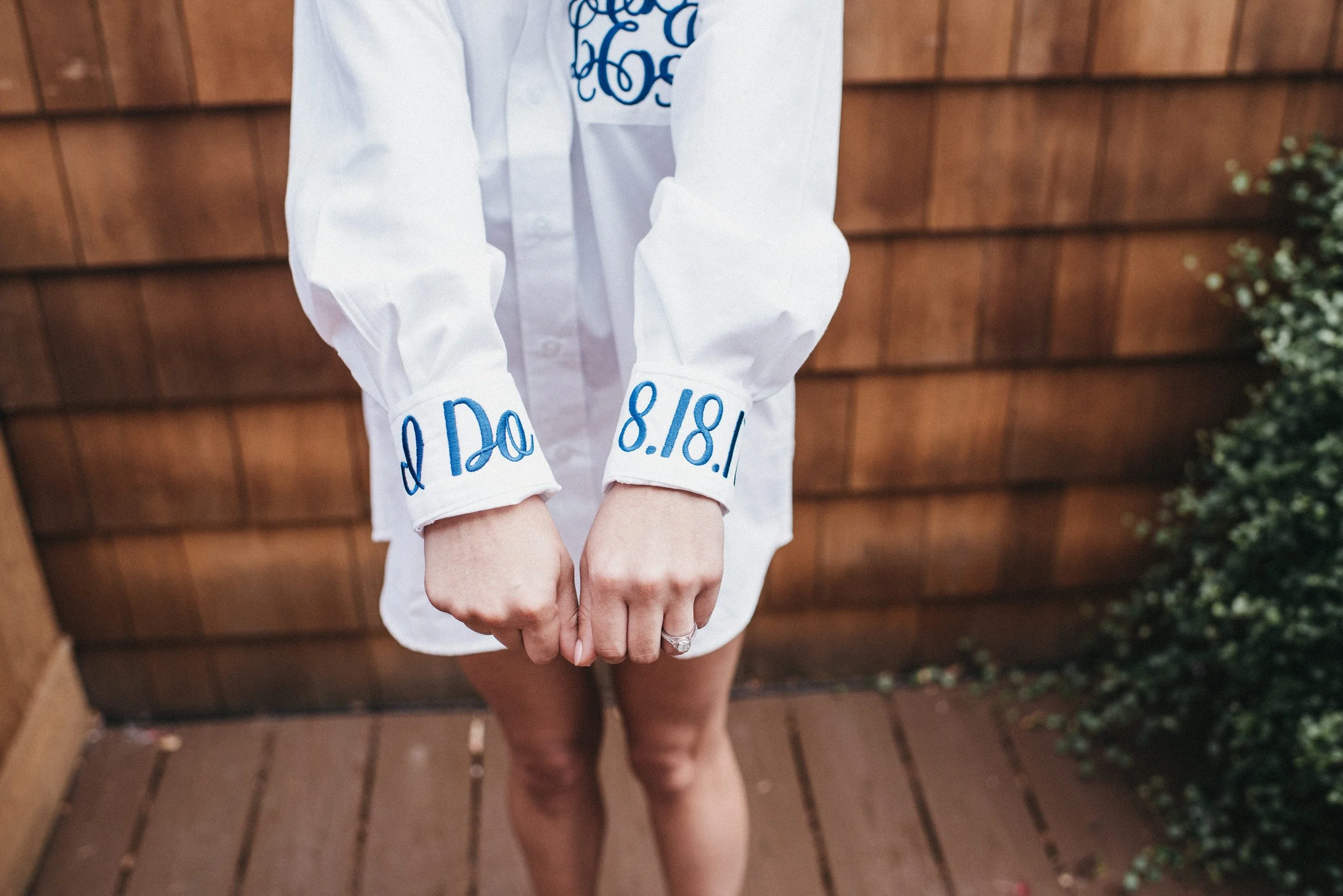 Person wearing a white shirt with blue embroidery and matching blue writing on the sleeves, standing on a wooden deck, with a wooden fence and greenery in the background.