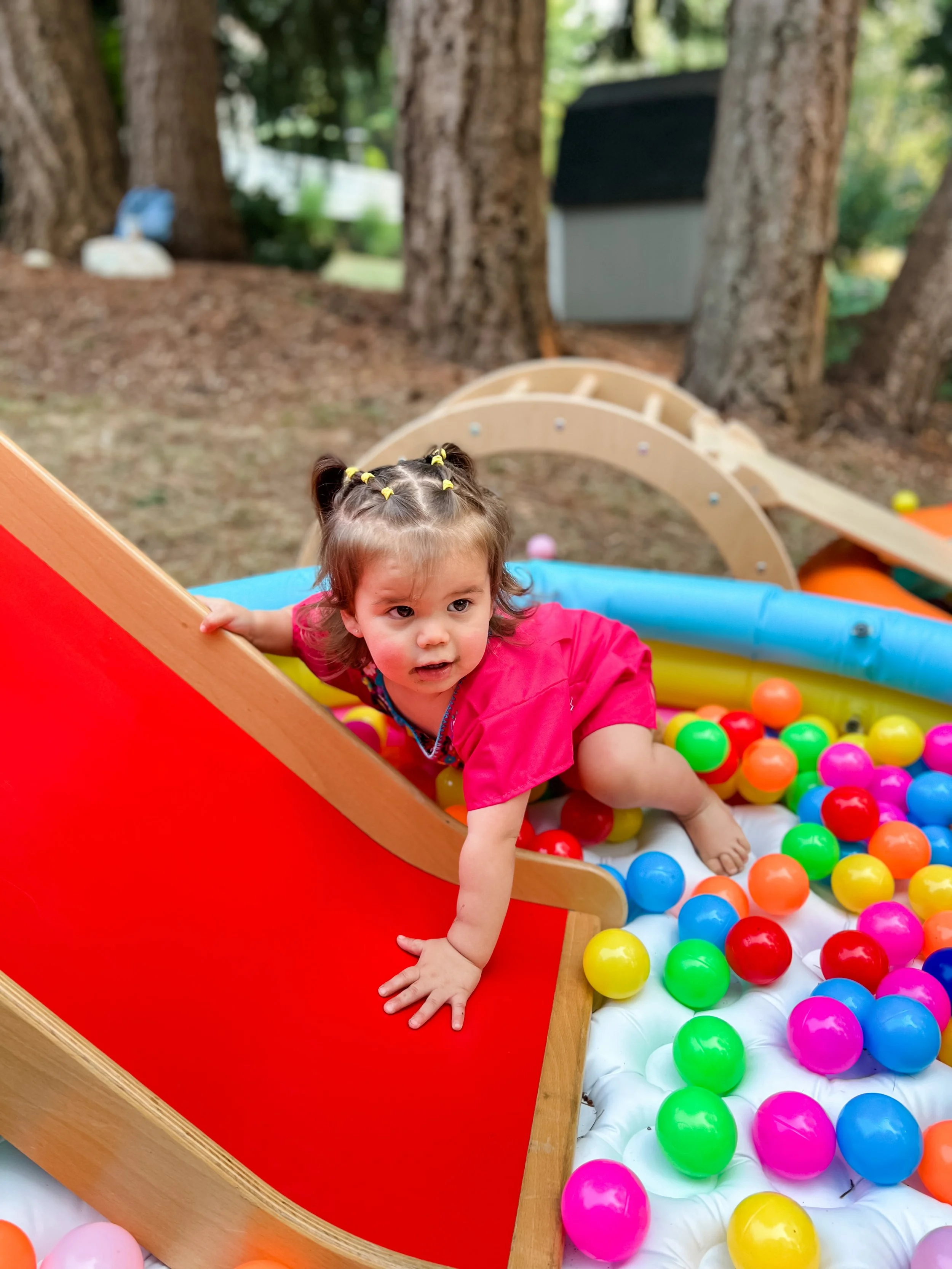 Young girl wearing a pink shirt on a colorful ball pit slide outdoors surrounded by trees.