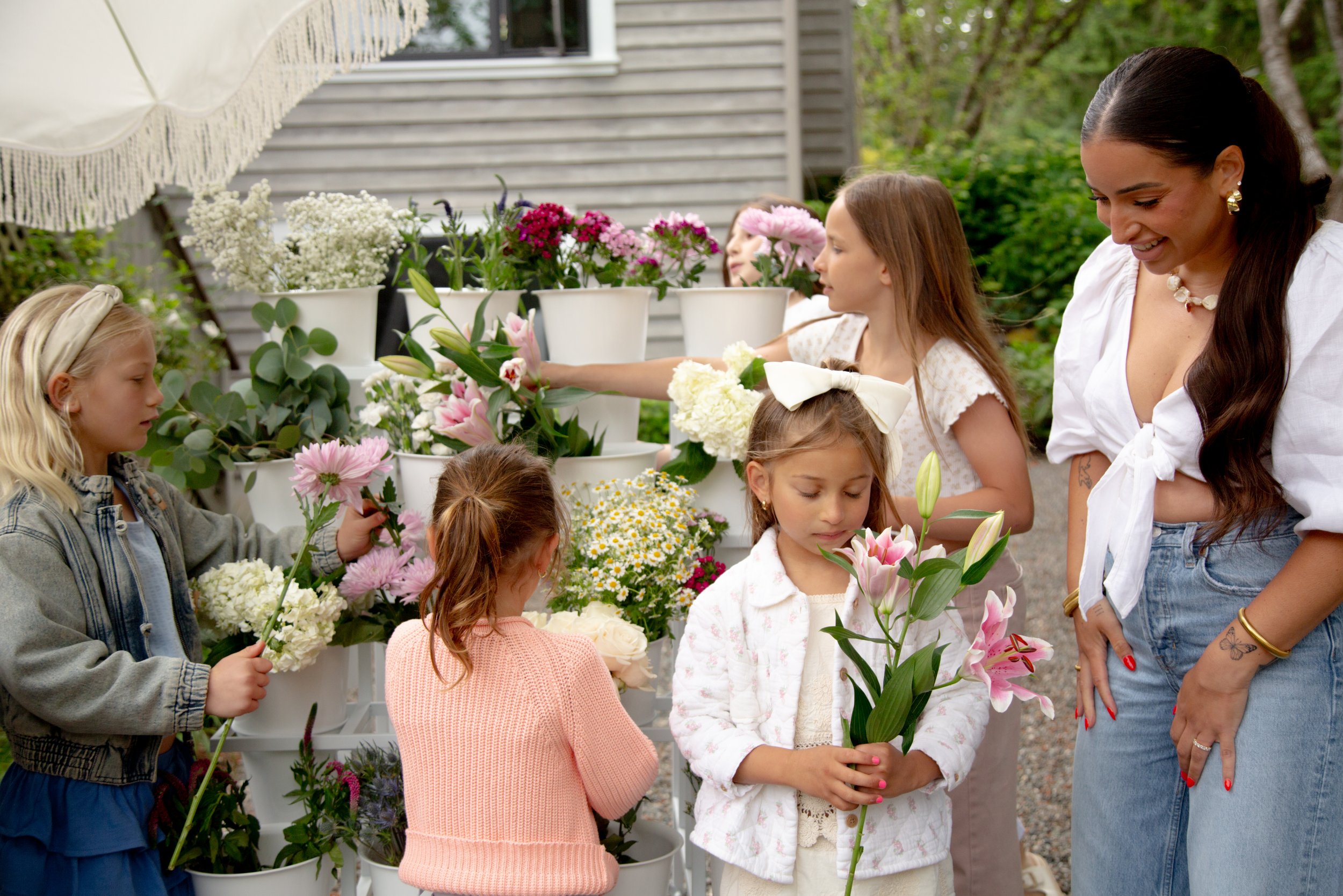 A group of children and an adult woman selecting flowers at an outdoor flower stand. The children are holding assorted flowers, and the woman is smiling. The background features potted flowers and greenery.