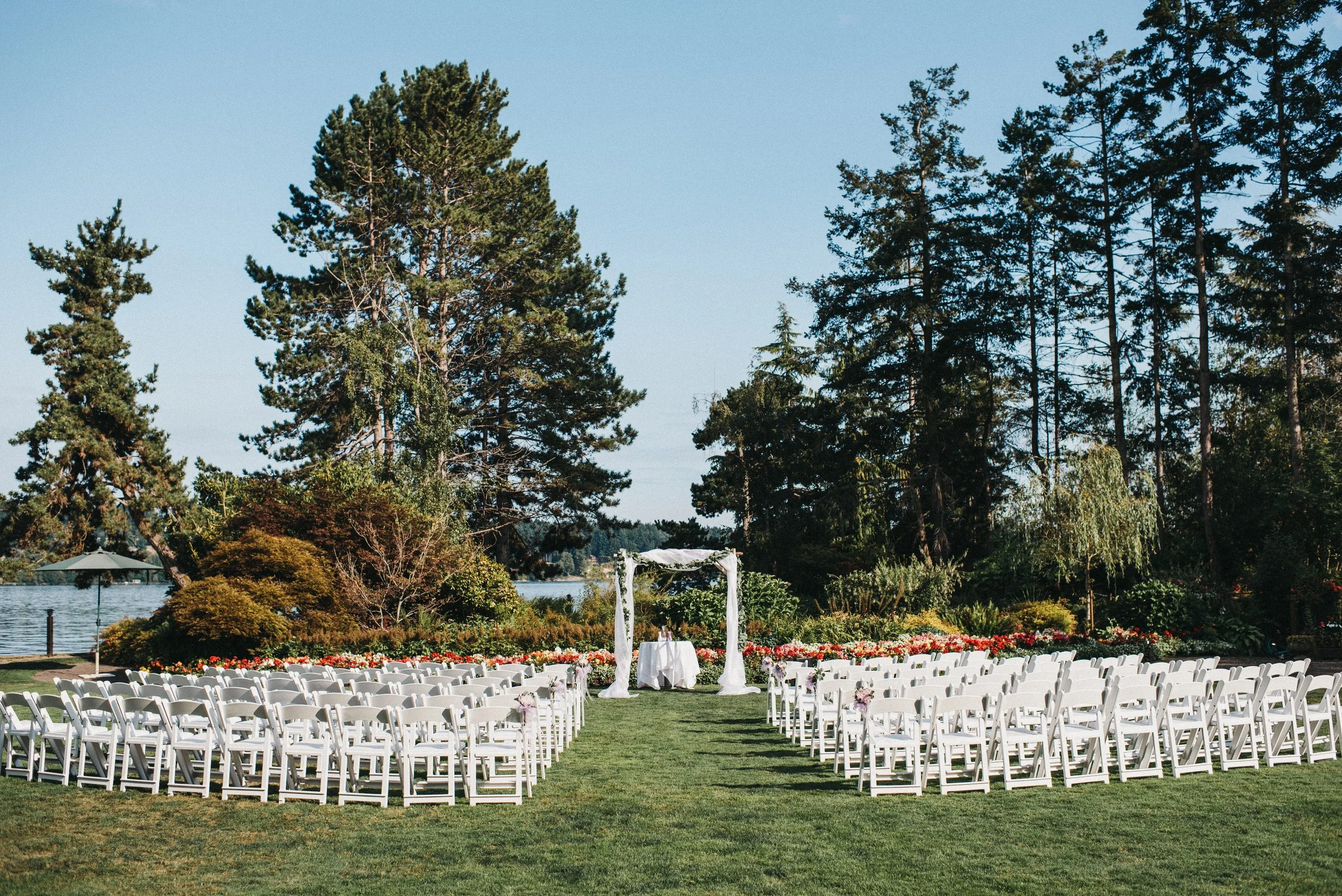 Outdoor wedding setup with white chairs arranged in rows on a grassy area facing a wedding arch. Trees and a lake are visible in the background under a clear blue sky.