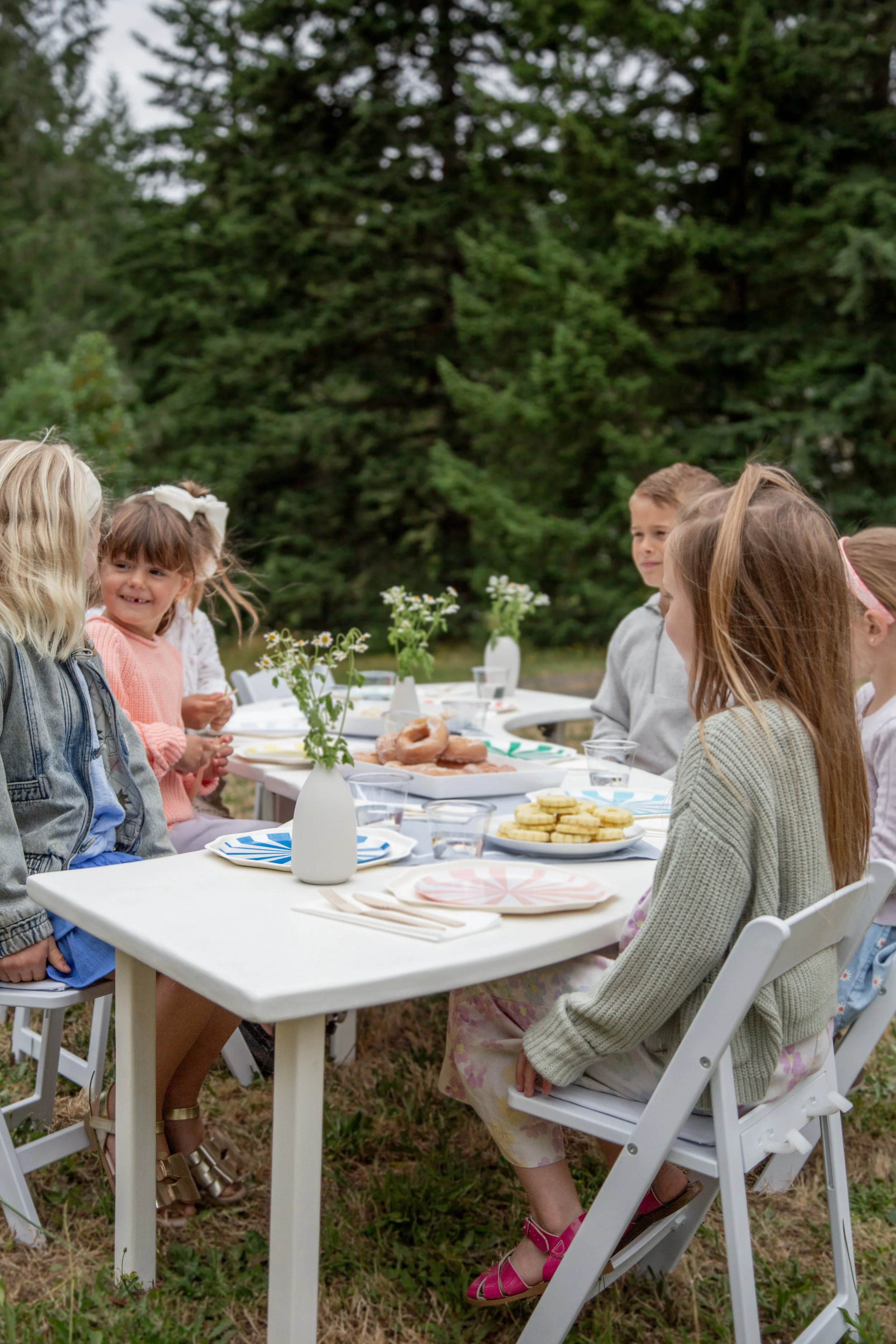 Children gathered around a table outdoors, celebrating with food and drinks, with a backdrop of green trees.