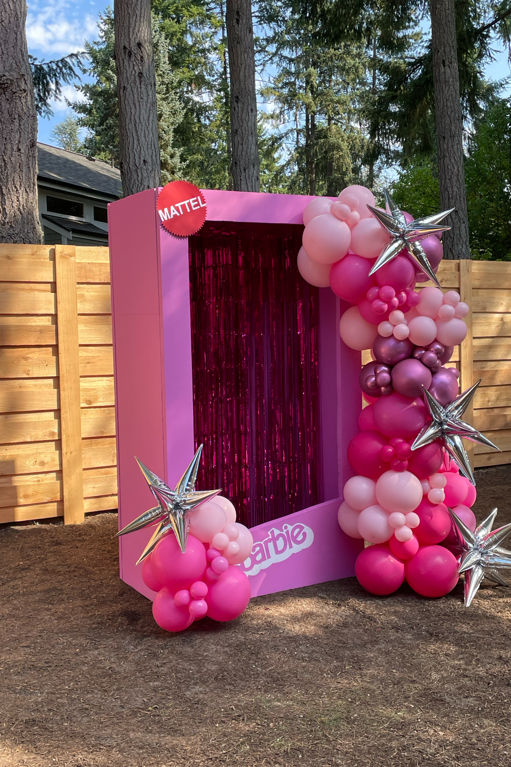 Decorative photo booth with a pink backdrop, surrounded by pink, purple, and silver balloons in various sizes, set outdoors with trees and a wooden fence in the background.