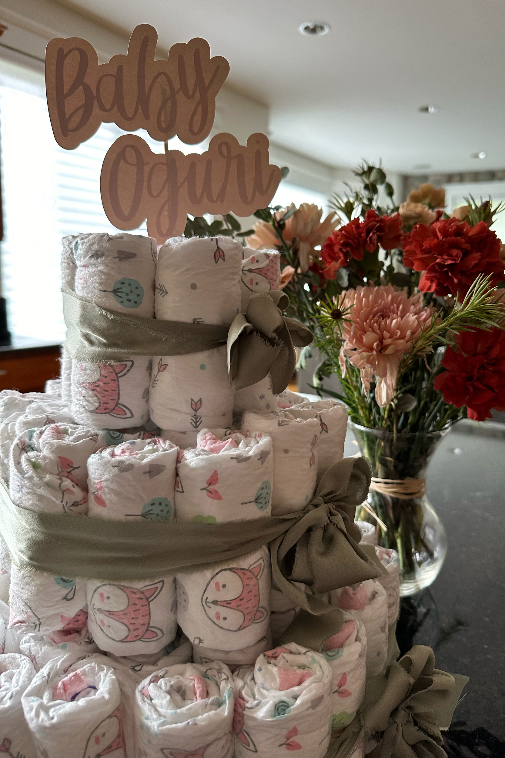 A table arrangement featuring a stack of newborn diapers with animal and nature patterns tied with a beige ribbon, a vase of pink and red flowers, and a sign that reads 'Baby On Board' in a child's handwriting style.