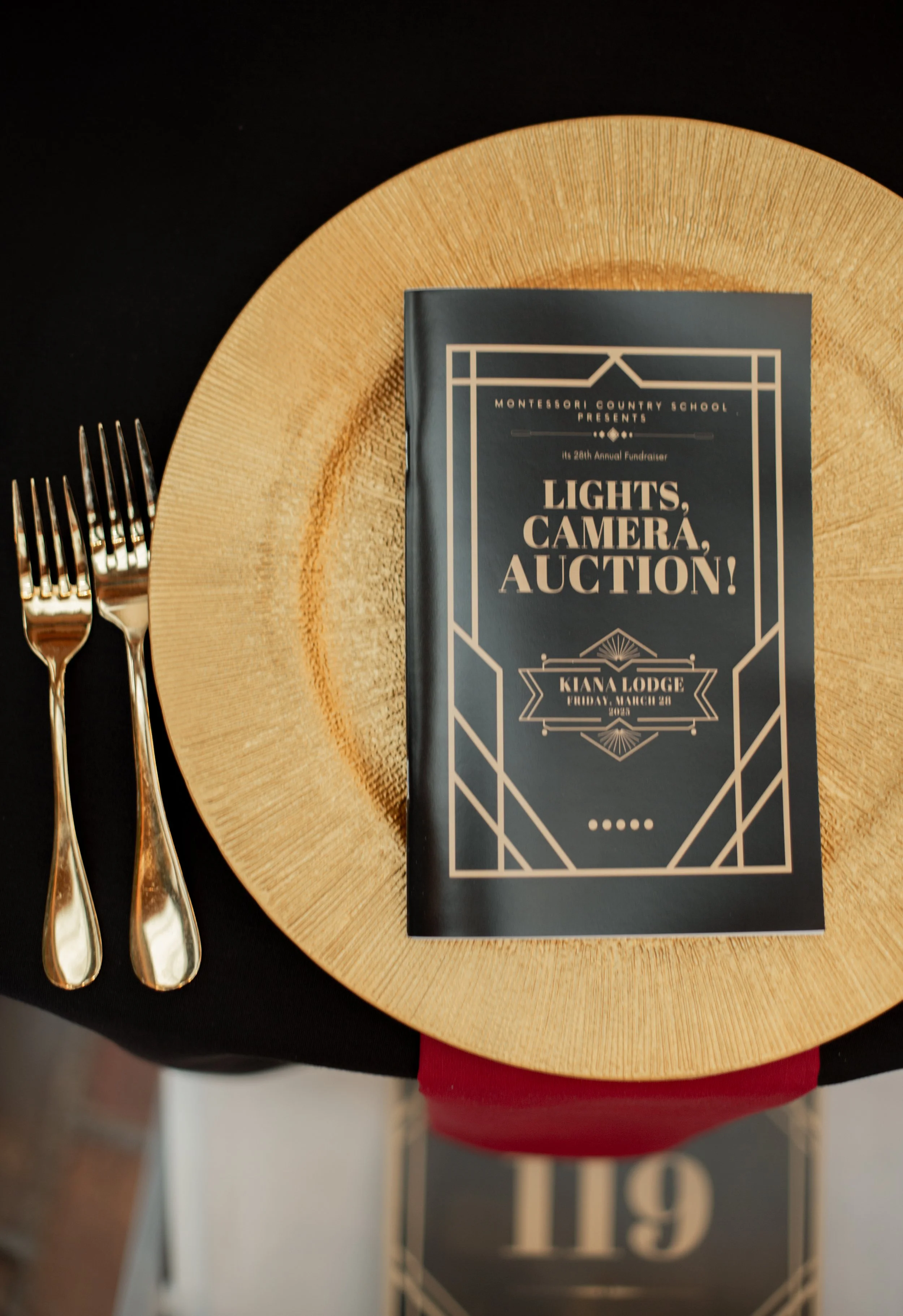 Event program for the 'Lights, Camera, Auction!' fundraiser at Kiara Lodge on Friday, March 20, 2020, placed on a round wooden charger plate with a black tablecloth, flanked by two silver forks.