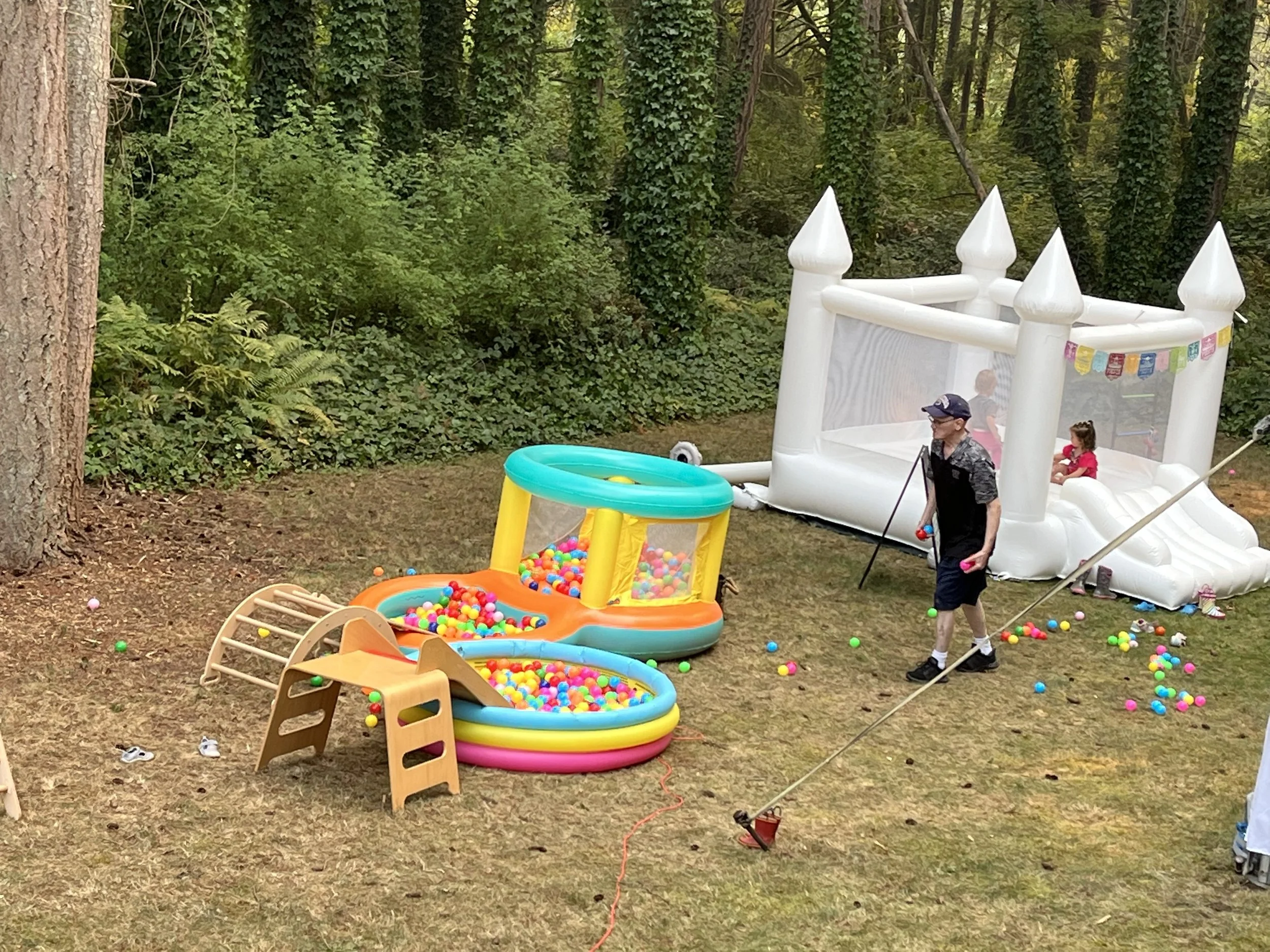 Children playing in inflatable castle and ball pit at an outdoor party in a wooded area.