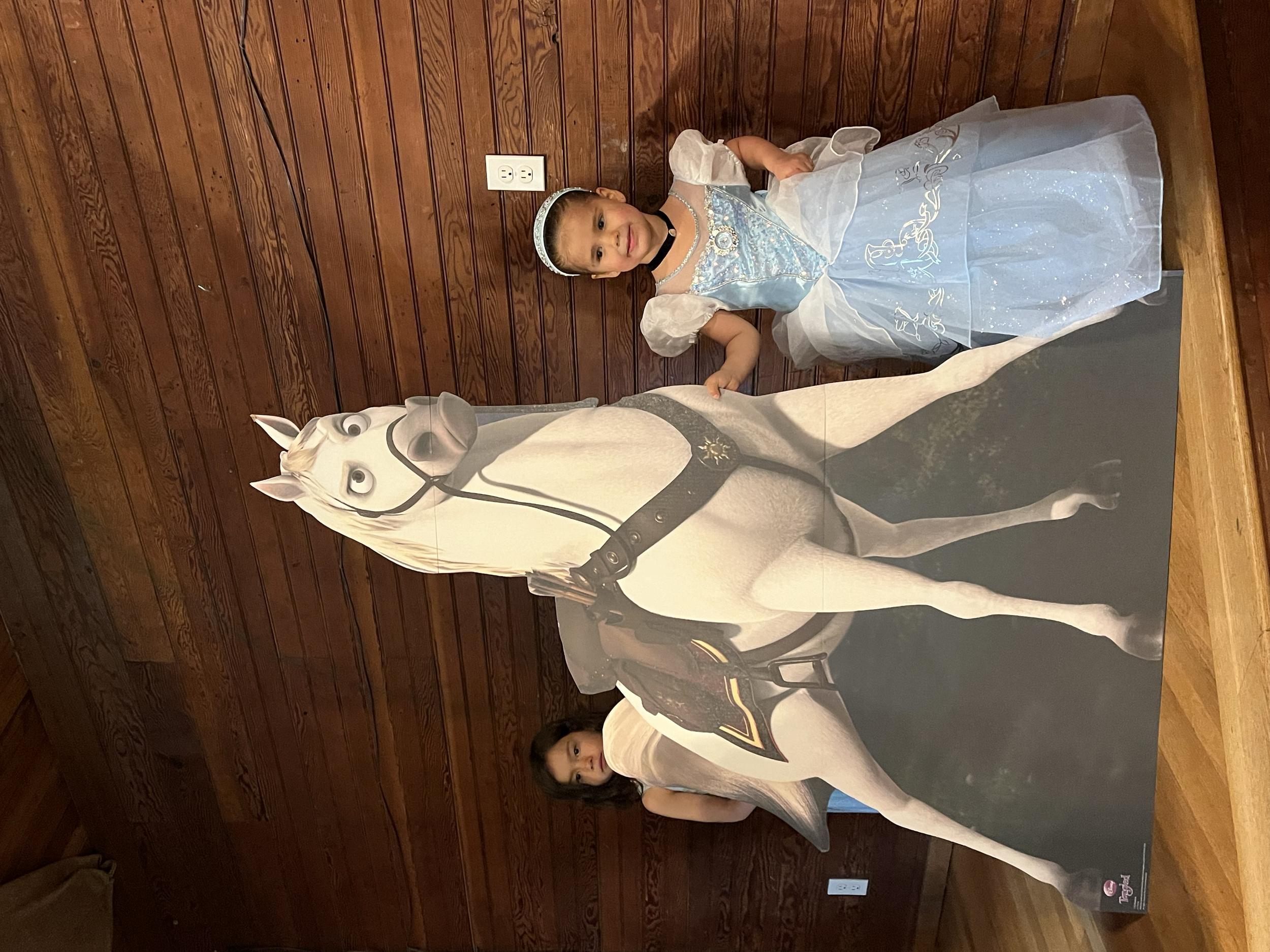 Two young girls stand behind an oversized cardboard cutout of a white horse with a girl's face on the horse's head, dressed as a princess for a costume event.