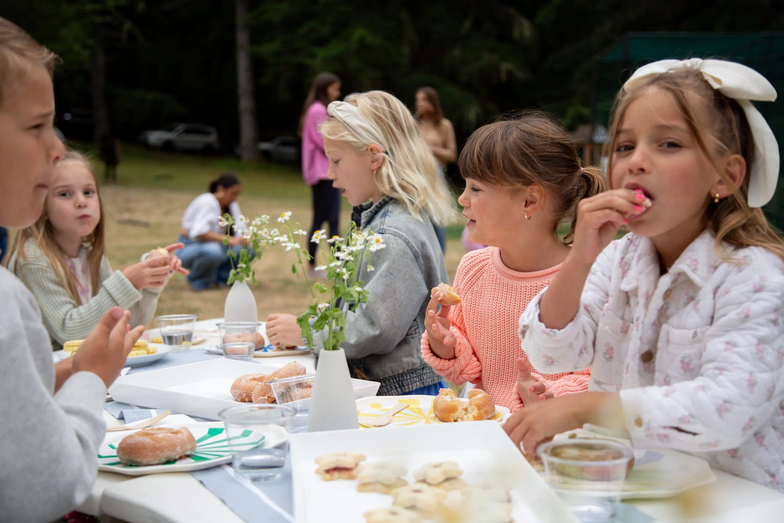 A group of children and adults enjoying a picnic outdoors with food and flowers, with trees in the background.
