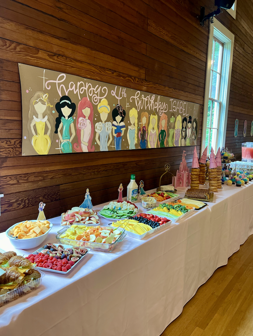 A dessert table with various fruits, pastries, and treats, decorated for a birthday party with a Disney princess theme, featuring a banner with cartoon princesses and stacked cookies resembling castle towers, set in a room with wooden walls and a lar