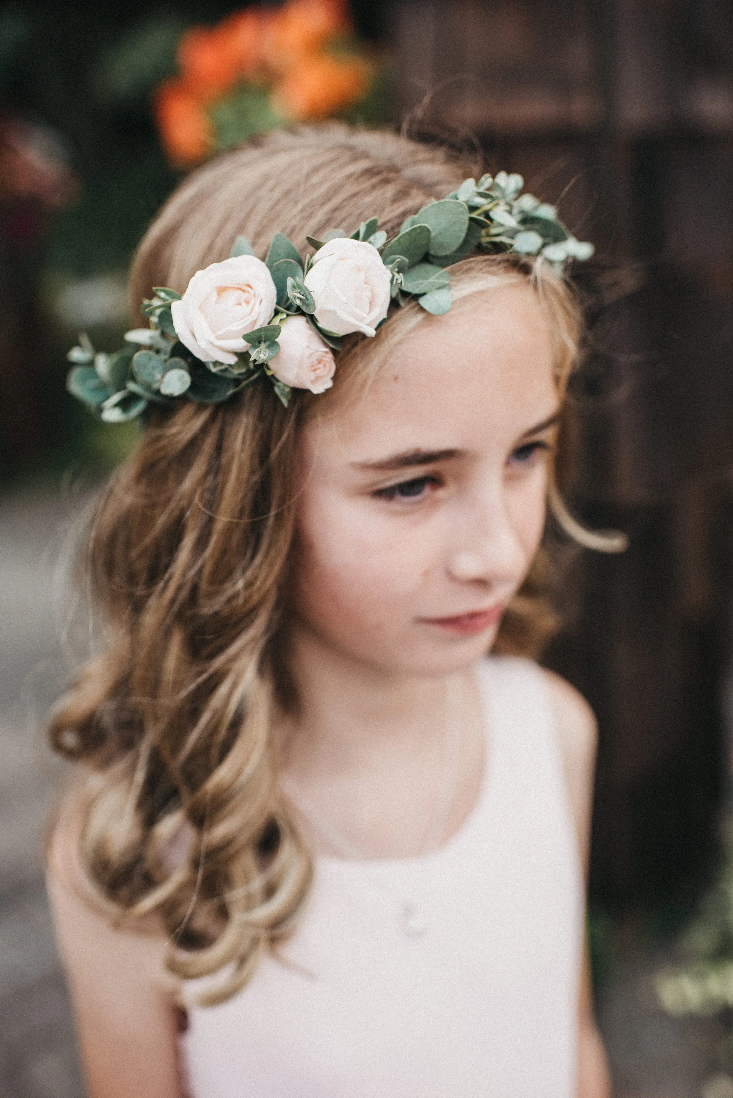 A young girl with light brown, curled hair wearing a white dress and a floral crown made of white roses and green leaves.