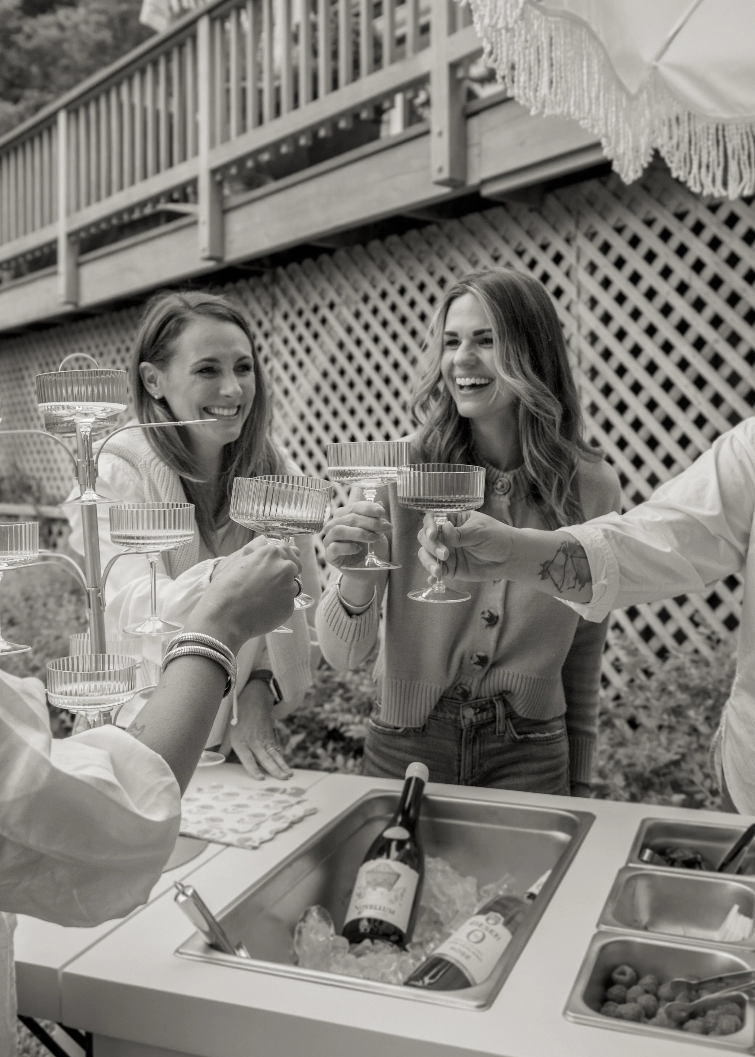Two women smiling and raising glasses at an outdoor gathering, with a third person partially visible. A table with various food items and drinks is in front of them, and a wooden deck with lattice and a patio umbrella are in the background.