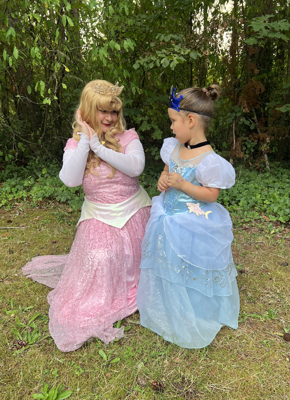 Two young girls dressed as princesses in costumes, one with a pink gown and crown, the other with a blue gown and a blue crown, standing outdoors on grass near trees.