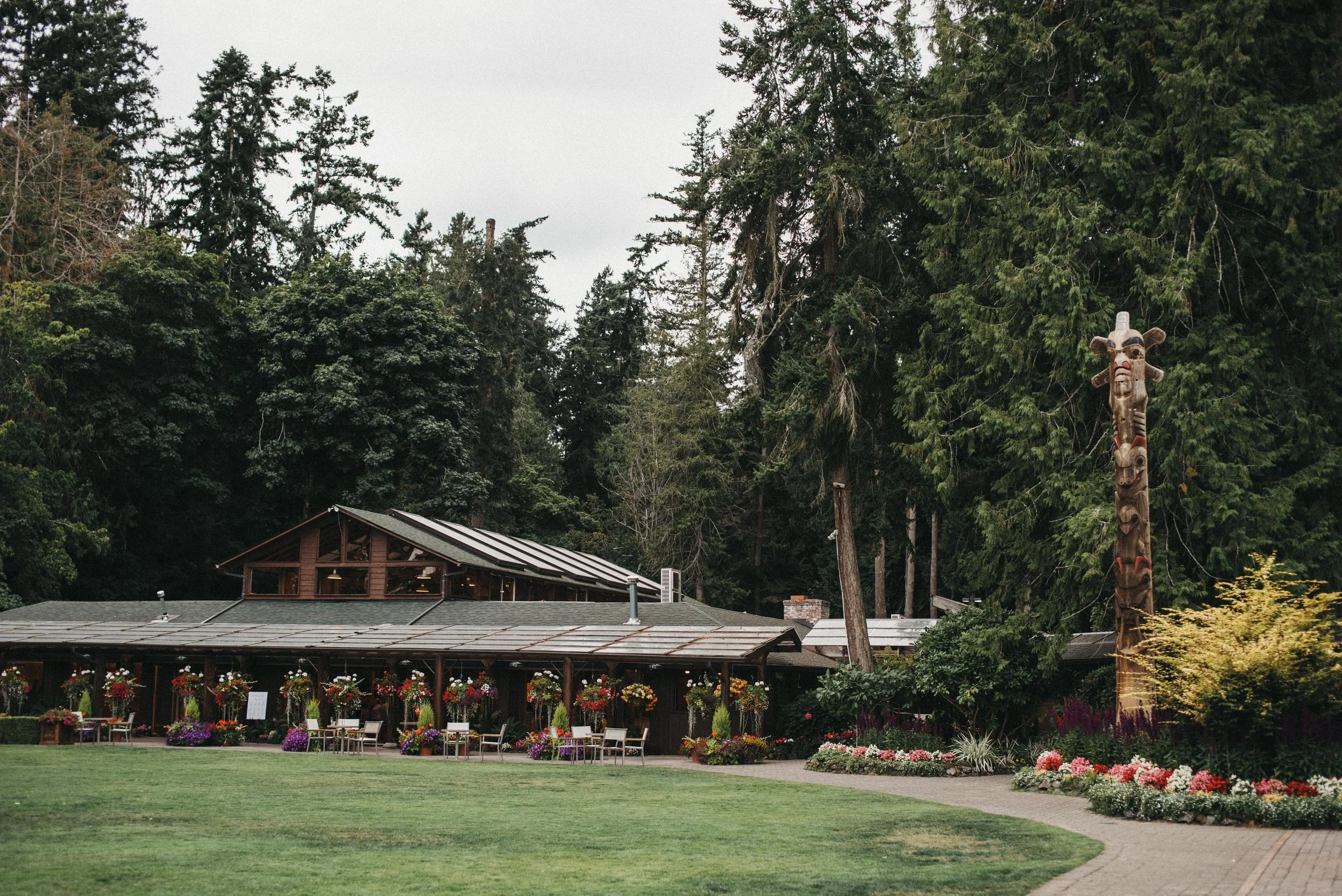 A building with a sloped roof surrounded by trees and colorful flowers, with a carved totem pole standing nearby.