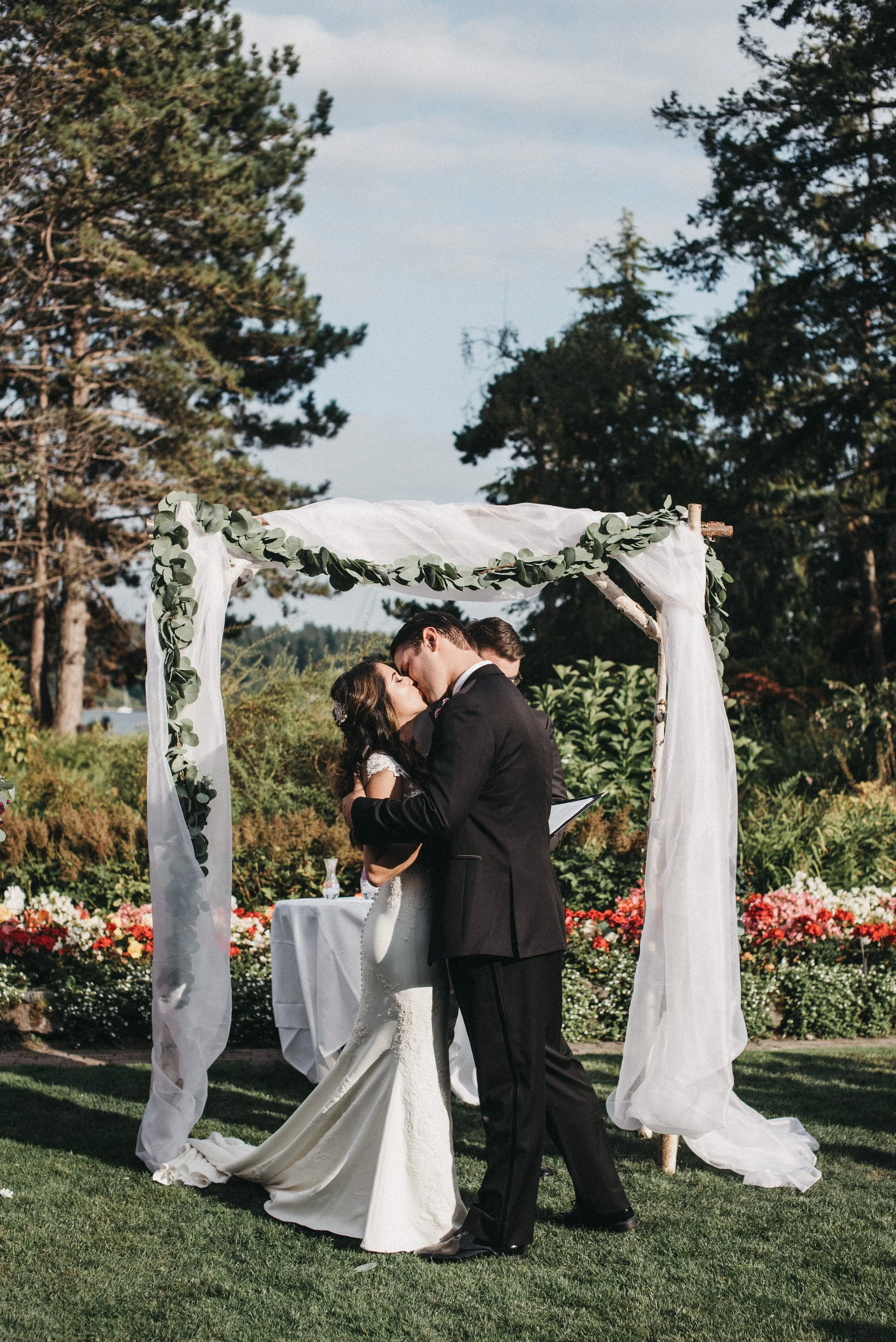 A couple in wedding attire sharing a kiss under an outdoor wedding arch decorated with greenery and white fabric, with a garden and trees in the background.