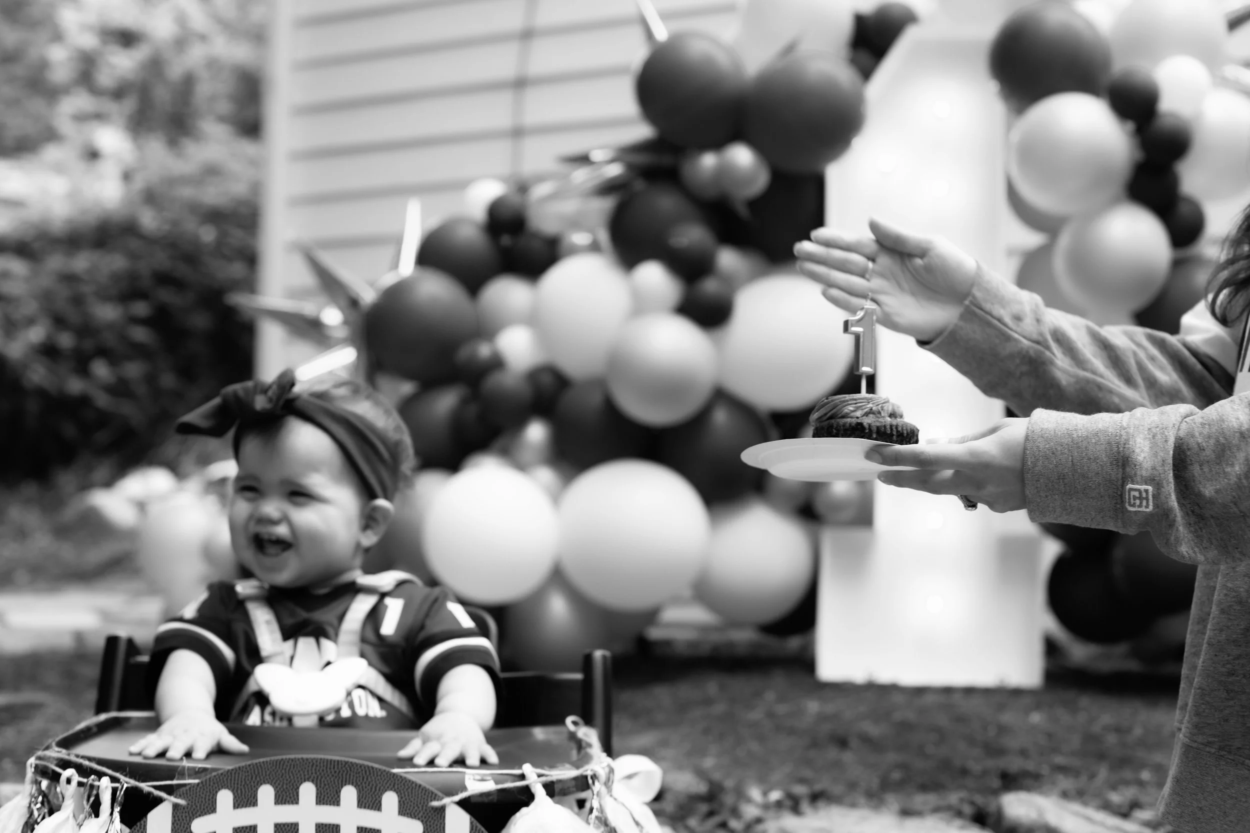 A baby in a stroller during a birthday celebration, smiling and laughing while a person holds a cupcake with a number one candle. In the background are balloons and a house siding.