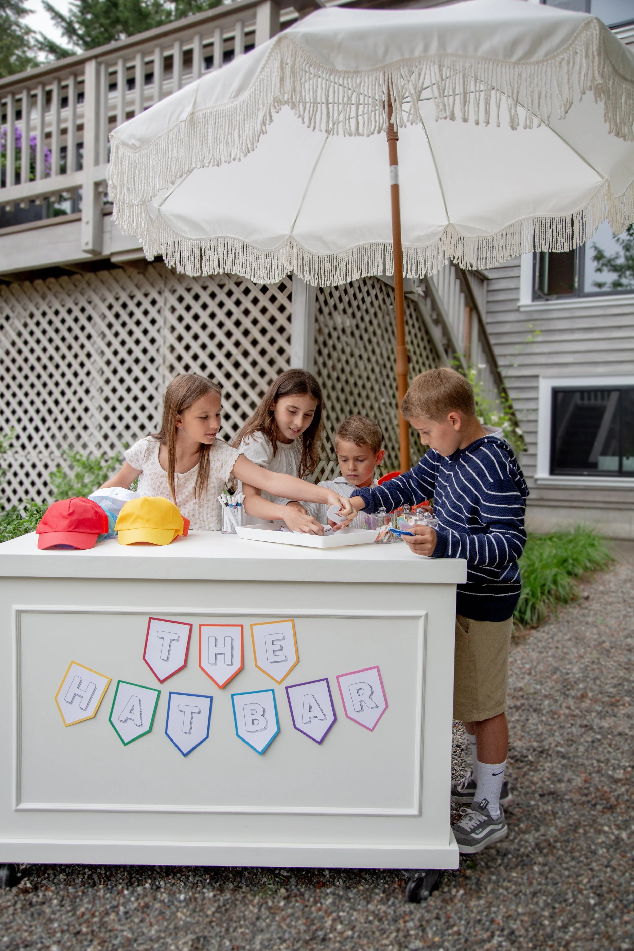 Children gathered around a craft booth at a yard sale, with a sign that says 'The Hat Bar' decorated with colorful flags.