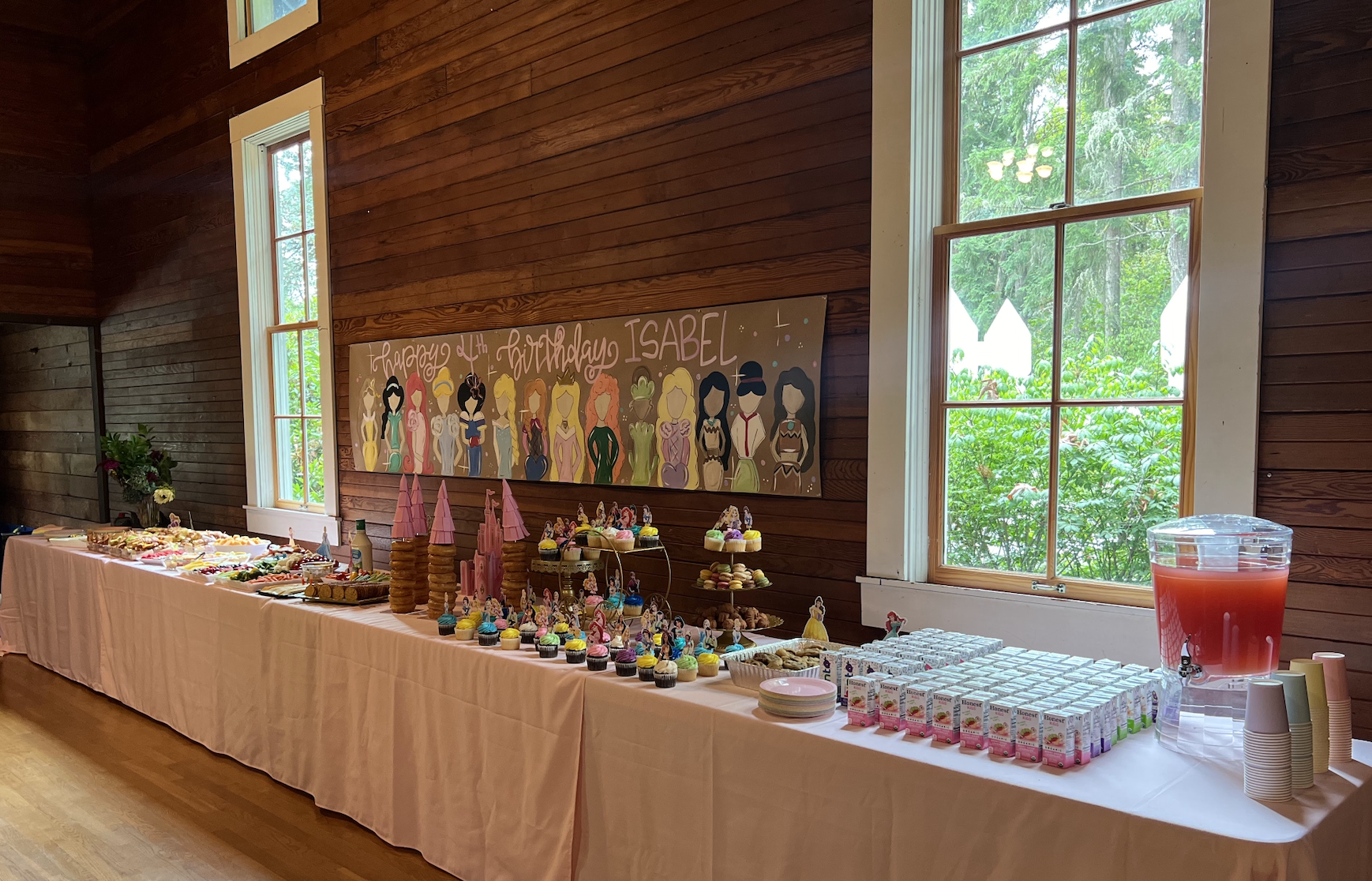 Dessert table decorated for a birthday party with cupcakes, cookies, and drinks in a rustic wooden room with large windows and a colorful birthday sign on the wall.