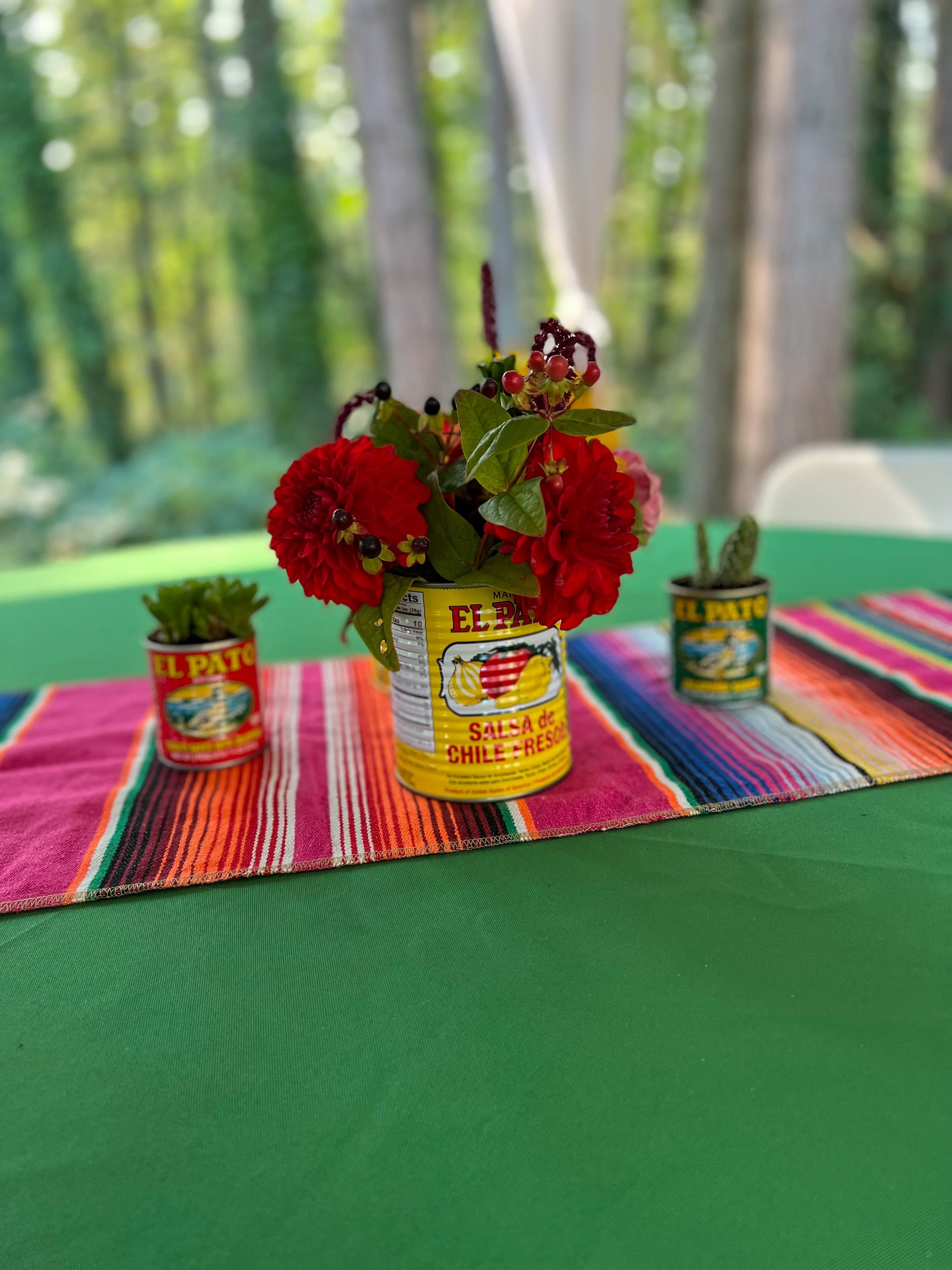 A fruit can decorated with red flowers and berries serving as a vase, placed on a colorful striped table runner on a green table, with two other cans containing plants on either side, set outdoors with trees in the background.