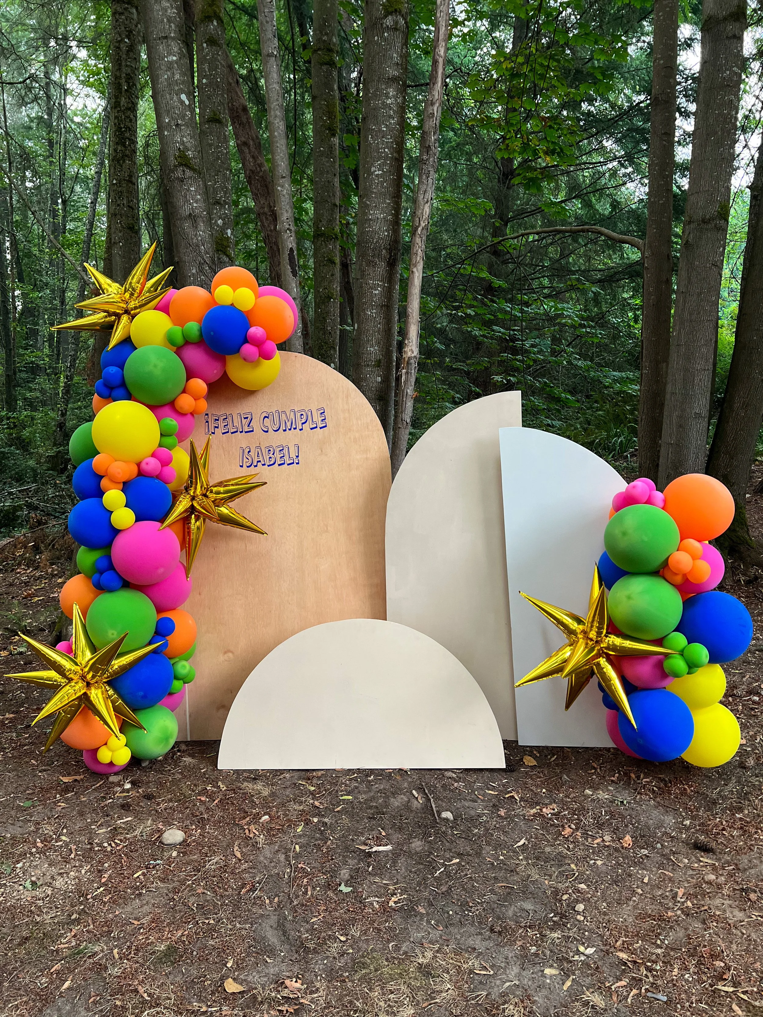 Colorful birthday photo backdrop decorated with balloons and metallic stars, with a message in Spanish that says 'Feliz Cumple Isabel!'. It is set outdoors among trees.