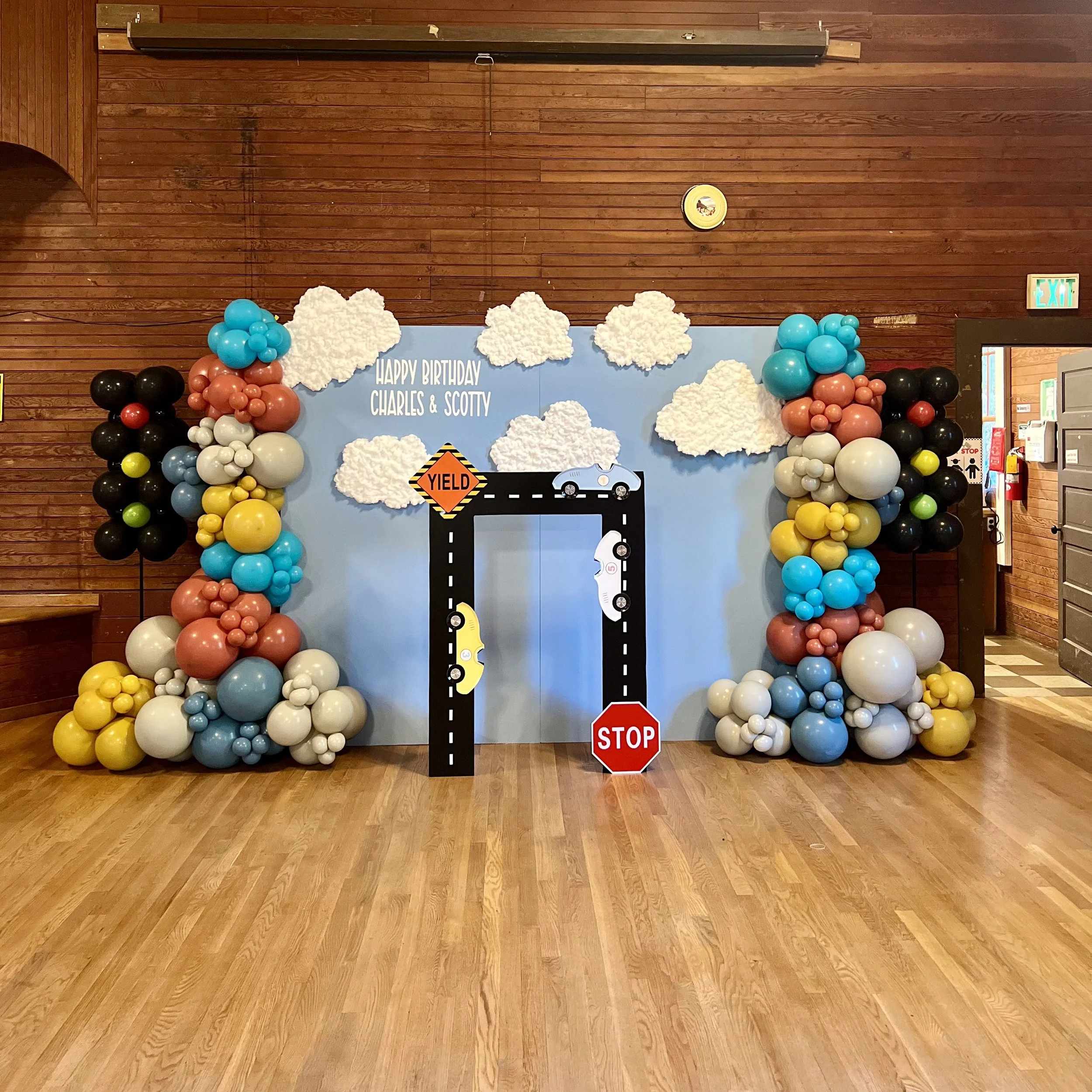 Birthday backdrop with a road theme, decorated with colorful balloons on the sides, white fluffy clouds, a YIELD sign, a STOP sign, and cartoon cars, with a message that reads 'Happy Birthday Charles & Scotty.'