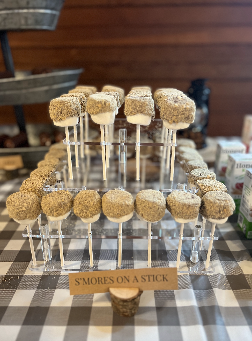 Sweet treats called s'mores on a stick, displayed in a tiered clear stand on a checked tablecloth.