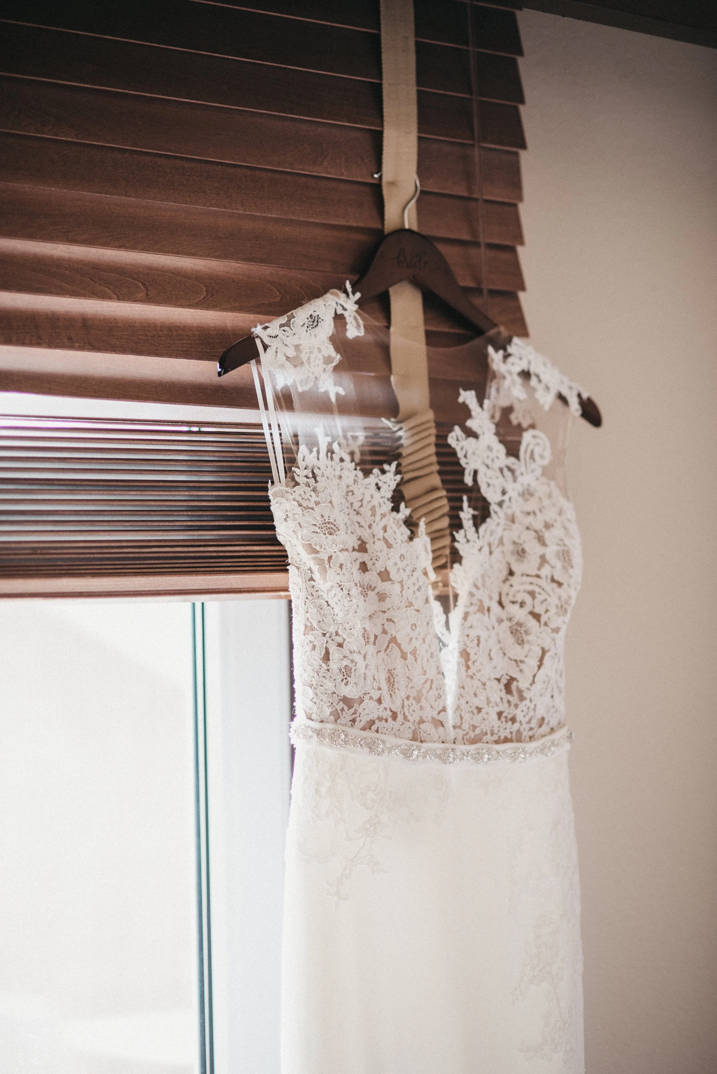 An elegant wedding dress with lace details, hanging on a wooden hanger near a window with brown blinds.