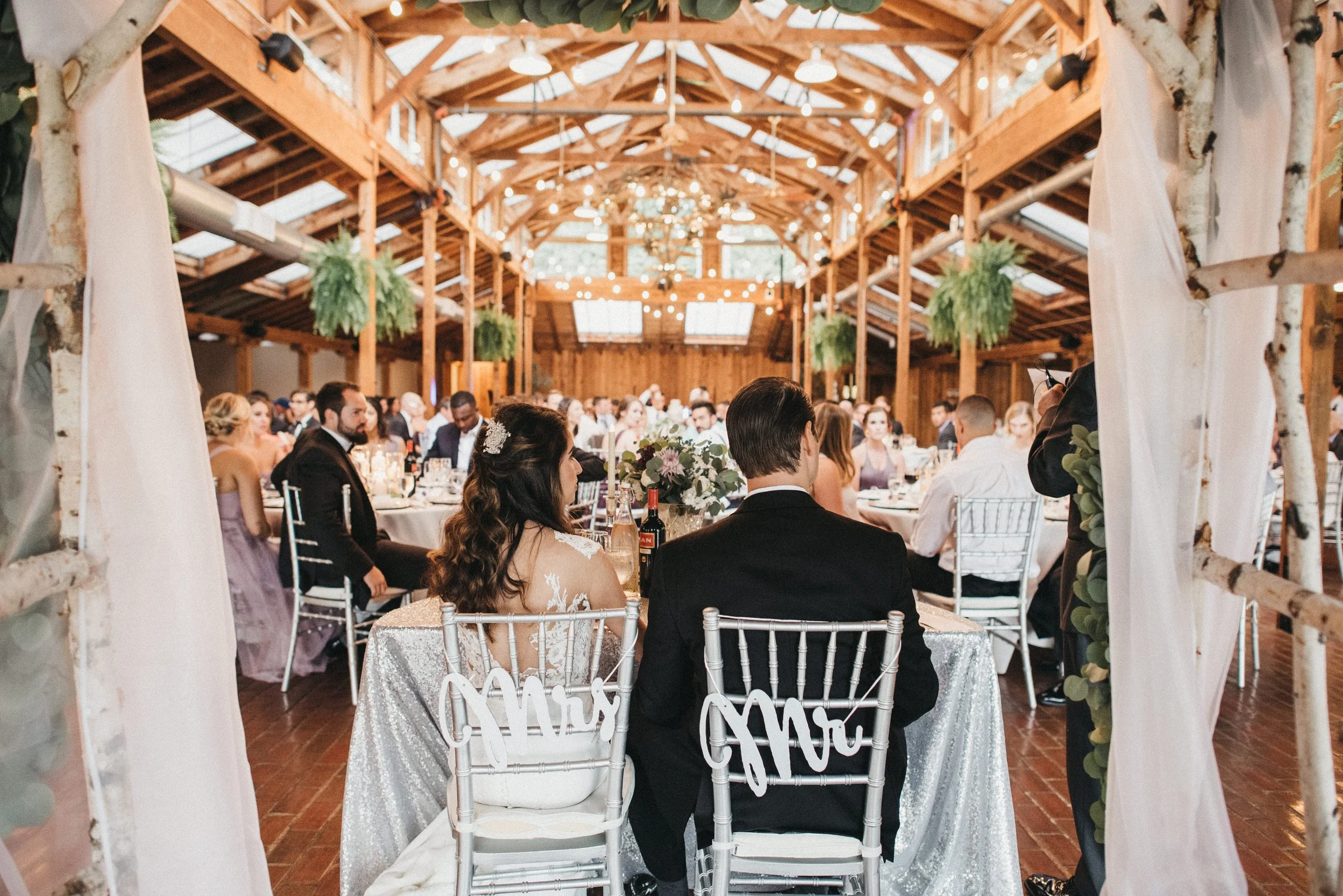 Wedding reception inside a rustic barn with wooden beams, string lights, and hanging greenery, featuring a bride and groom seated at a decorated table with a crowd of guests in the background.