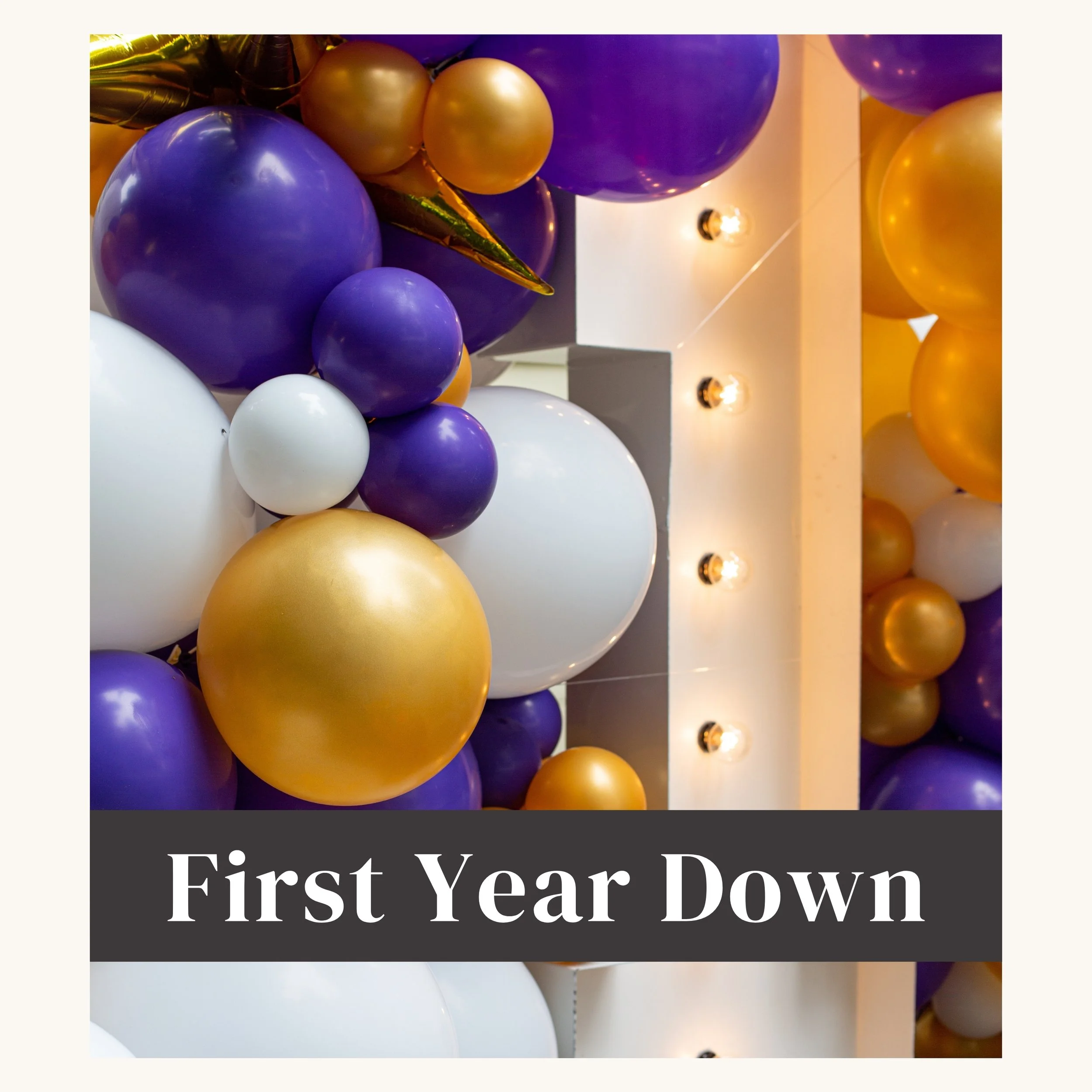 Colorful balloons in purple, gold, and white with string lights and a sign that reads "First Year Down".