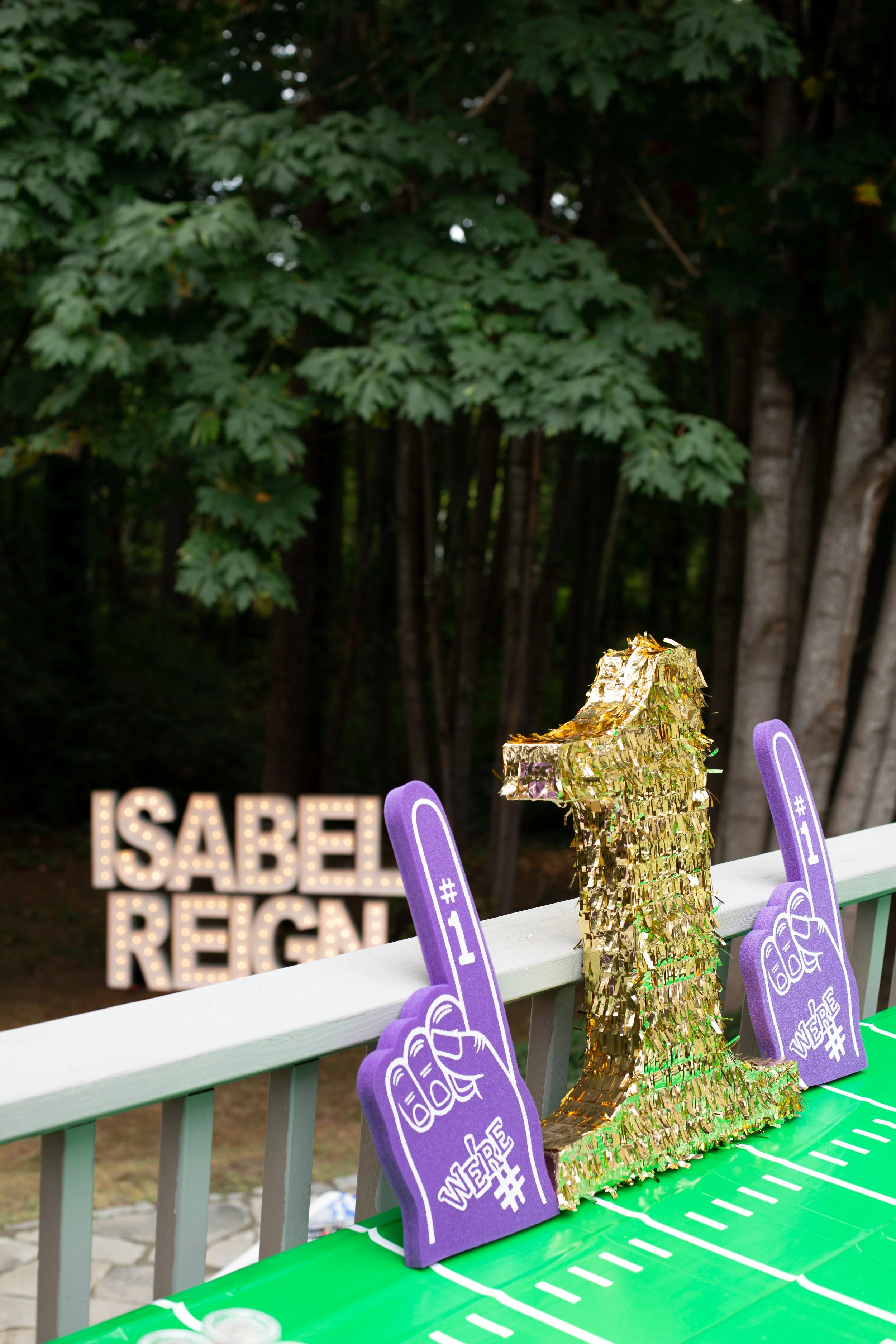 Decorative setup with purple foam fingers and a gold inflatable lion for celebrating Isabel's reign. A blurred illuminated sign in the background reads 'ISABEL REIGN.'