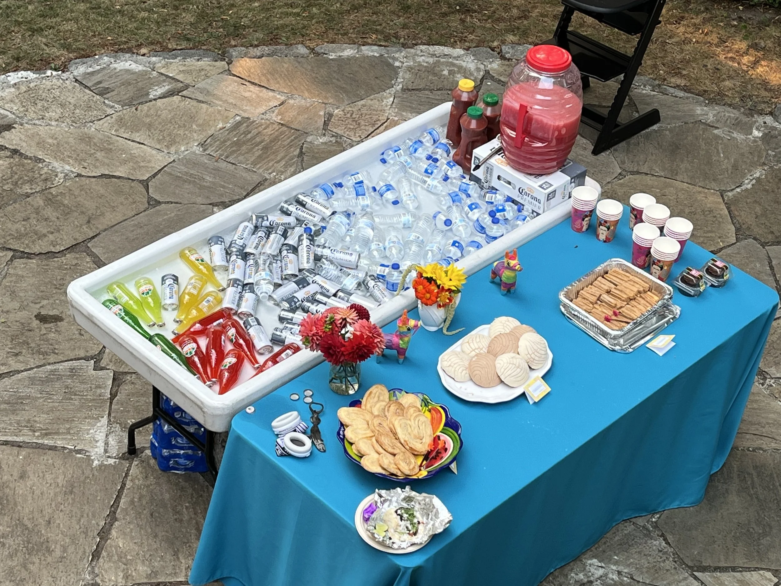 A table set up outdoors with various snacks and drinks, including bottled waters, colorful drinks, cookies, crackers, and chips, along with paper cups, flowers, and party decorations, on a blue tablecloth on a stone patio.