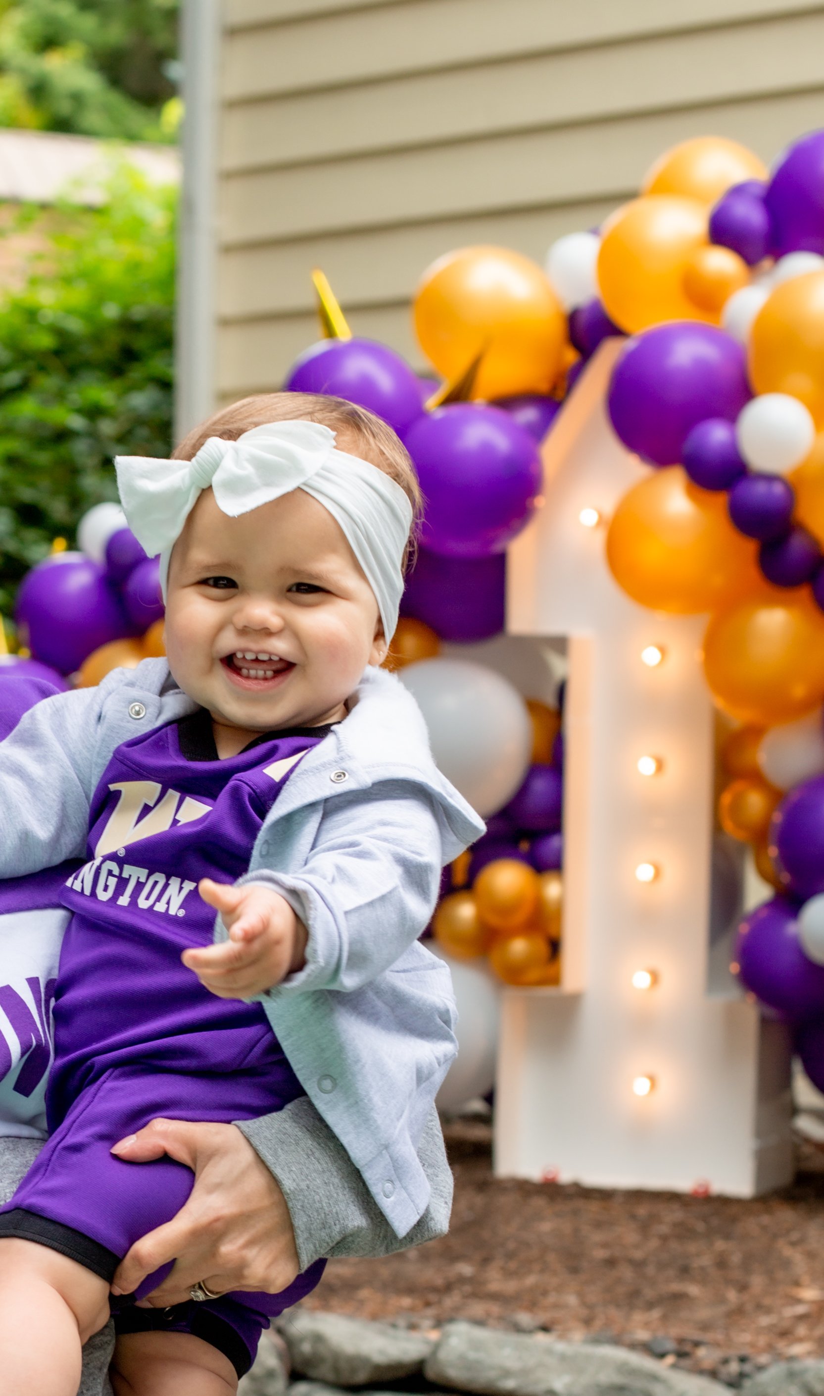 A smiling toddler girl with a white bow headband, wearing a purple sports jersey, held up in front of a decorative backdrop of orange, purple, white balloons, and a lit large white letter 'L' at an outdoor celebration.