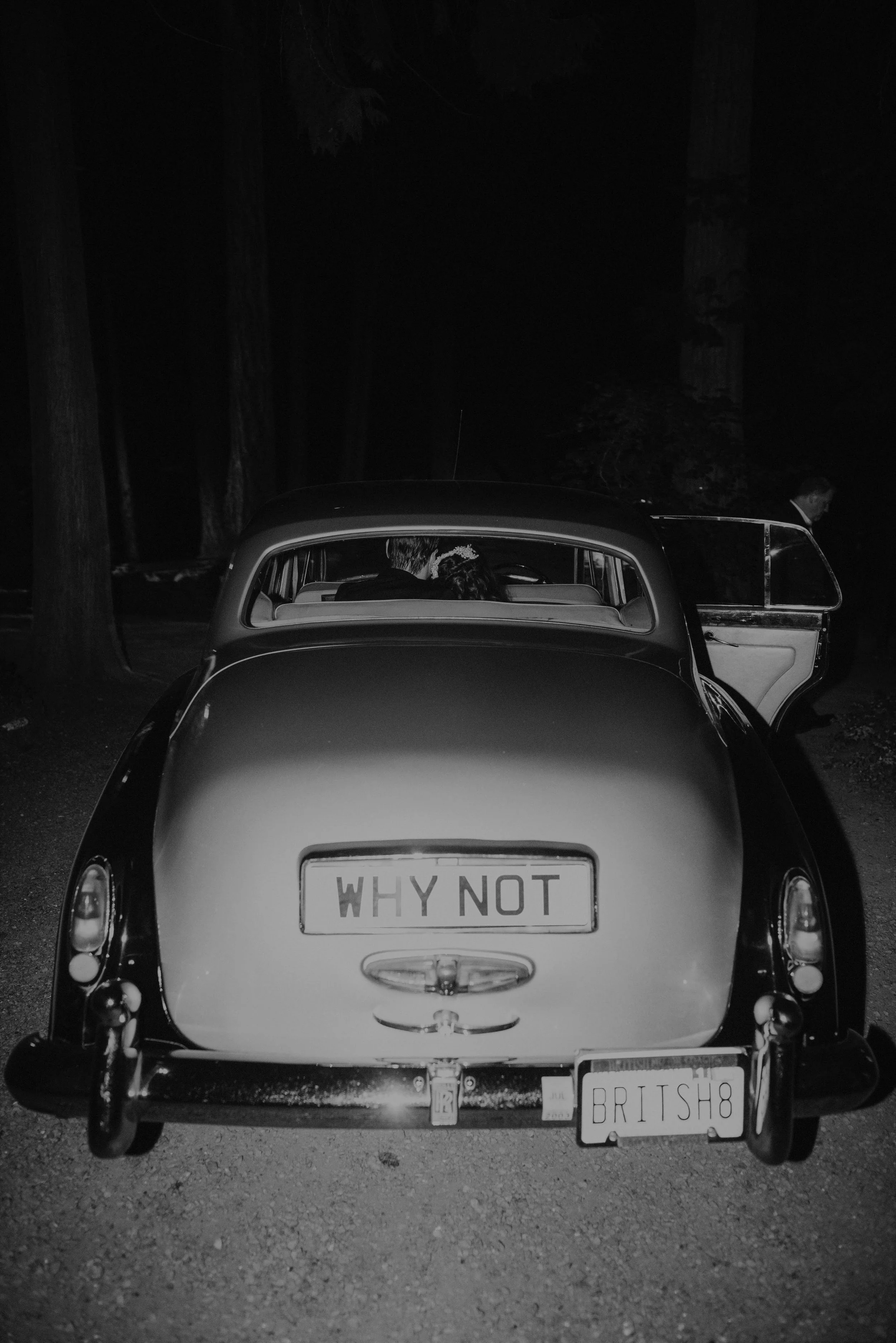 A vintage car parked outdoors at night, with a license plate that reads "WHY NOT" and a second license plate reading "BRITSH8." The car's rear window shows two people inside, and a man is seen standing near the open door on the right side of the imag