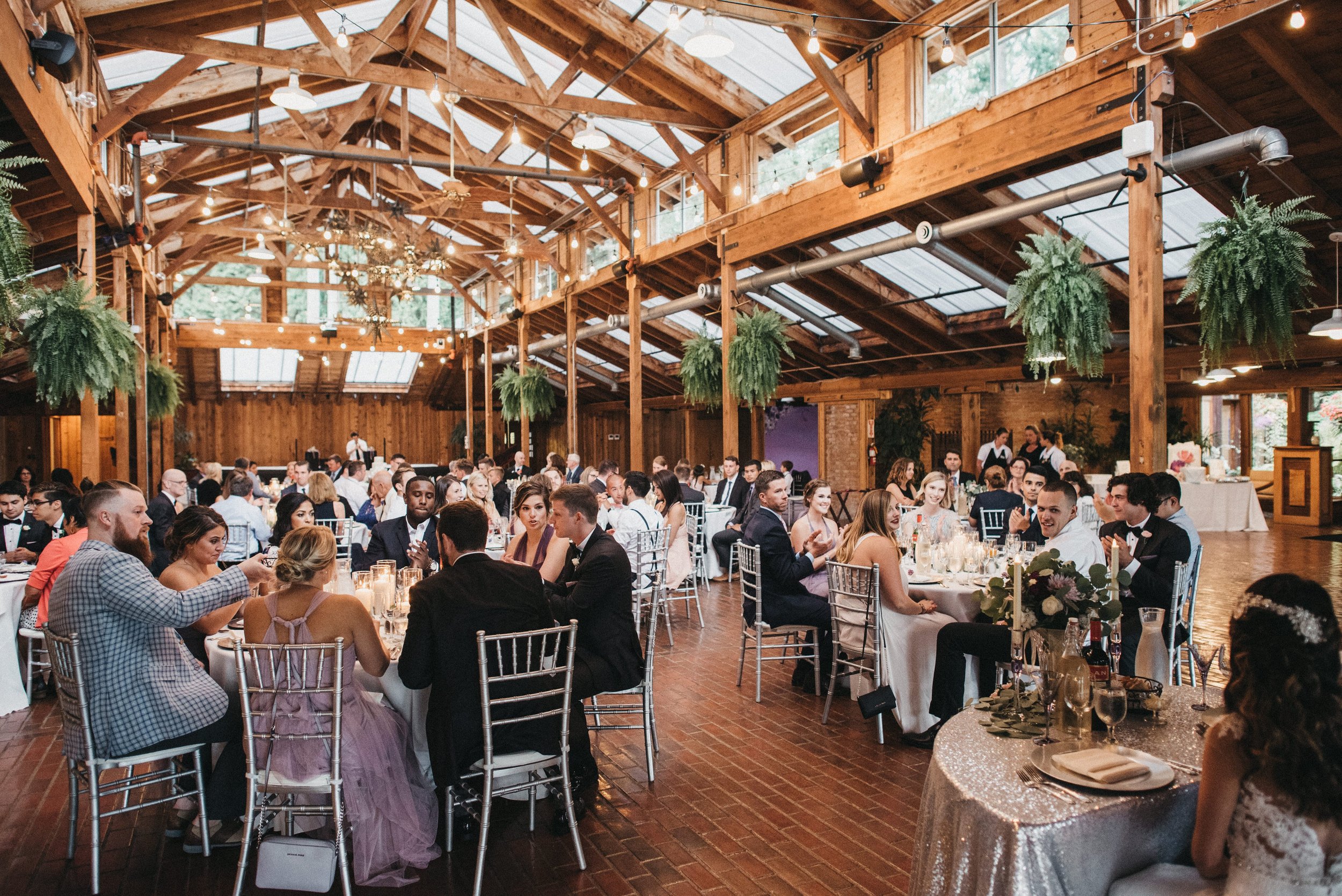 Guests seated at tables during a wedding reception in a rustic, wooden venue with hanging plants and string lights.