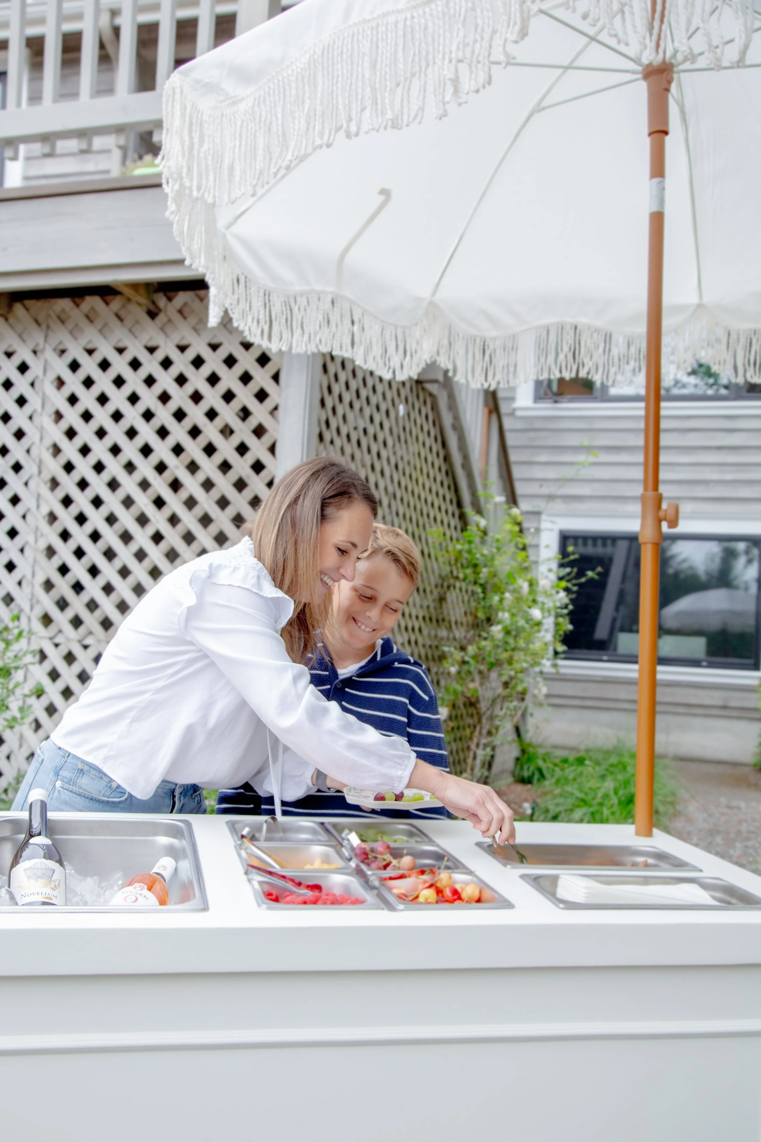 A woman and a young boy preparing food outdoors at a white kitchen island with various fresh fruits, with a white patio umbrella overhead and a wooden house in the background.